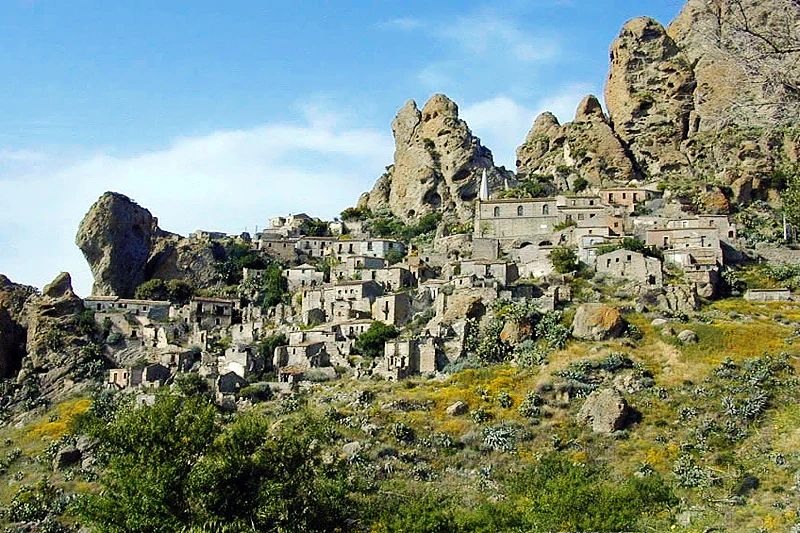 Stone village buildings on a rocky hillside with large rock formations and greenery under a clear blue sky