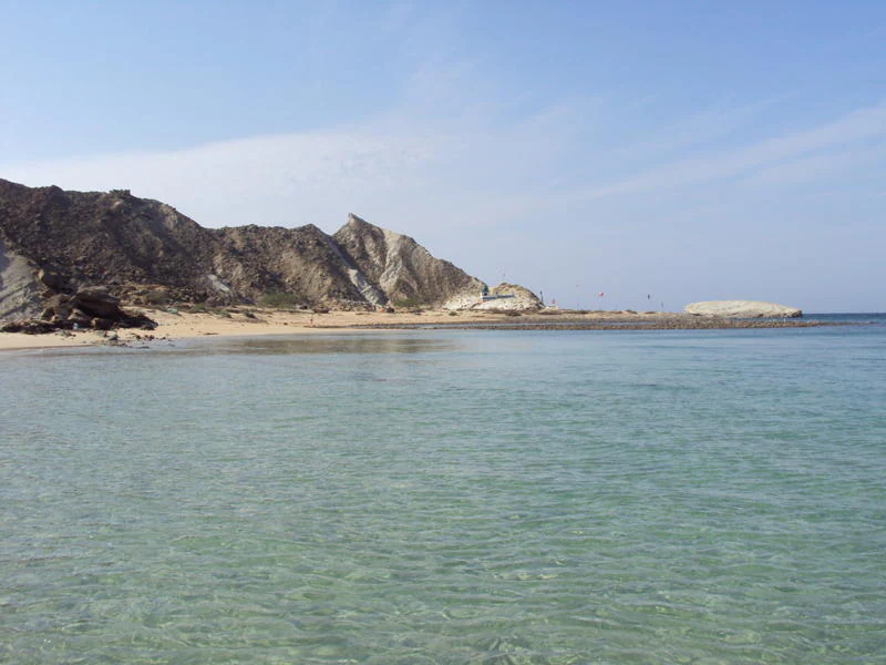 A wide angle view of the rugged, rocky coastline of Astola Island meeting clear, shallow turquoise water under a bright blue sky.