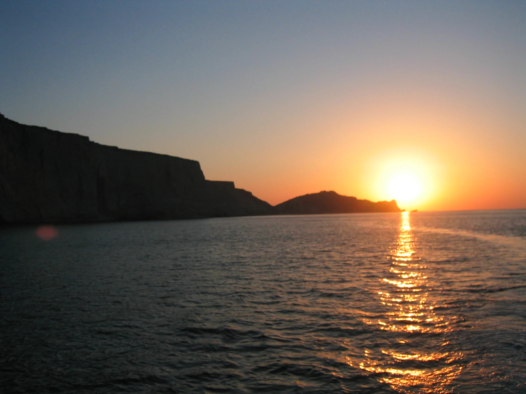 A wide view of the ocean at sunset with the dark silhouette of the cliffs of Astola Island in the background.