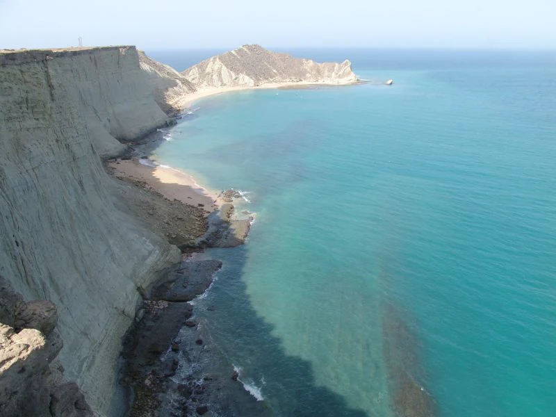 An elevated, wide-angle view of a rocky coastline in Astola Island with steep, light-colored cliffs dropping directly into clear, bright turquoise sea water.