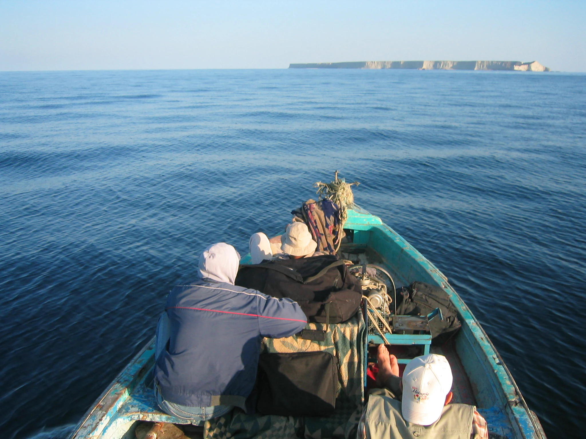 A view from the back of a small motorboat traveling on calm, blue water toward a flat, rocky island in the distant horizon.