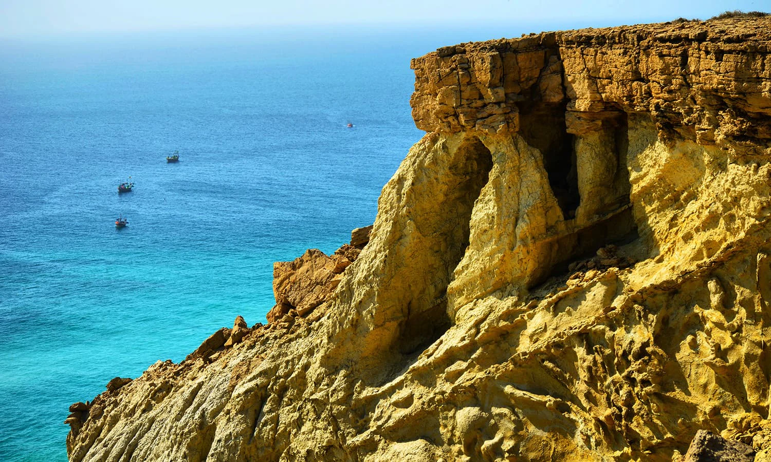 A high-angle view showing a jagged, sunlit rocky cliff face falling into deep blue and turquoise ocean water, with small fishing boats visible in the distance.