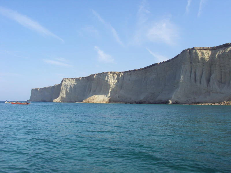 A wide view of the steep, light-colored limestone cliffs of Astola Island meeting the blue sea, with a small boat visible in the water near the base.