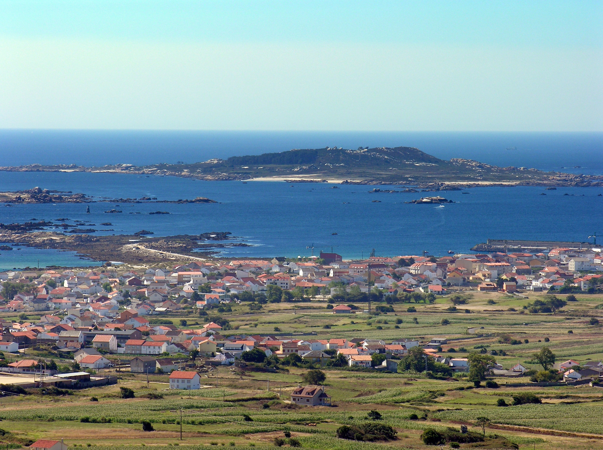 Coastal landscape showing Sálvora Island in the distance, with Aguiño village and surrounding fields in the foreground, overlooking the sea under a clear sky