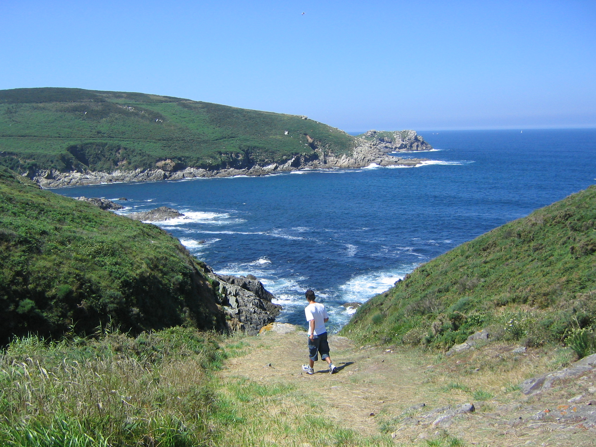 A person standing on a grassy path leading to a coastal bay with rocky cliffs and blue water.