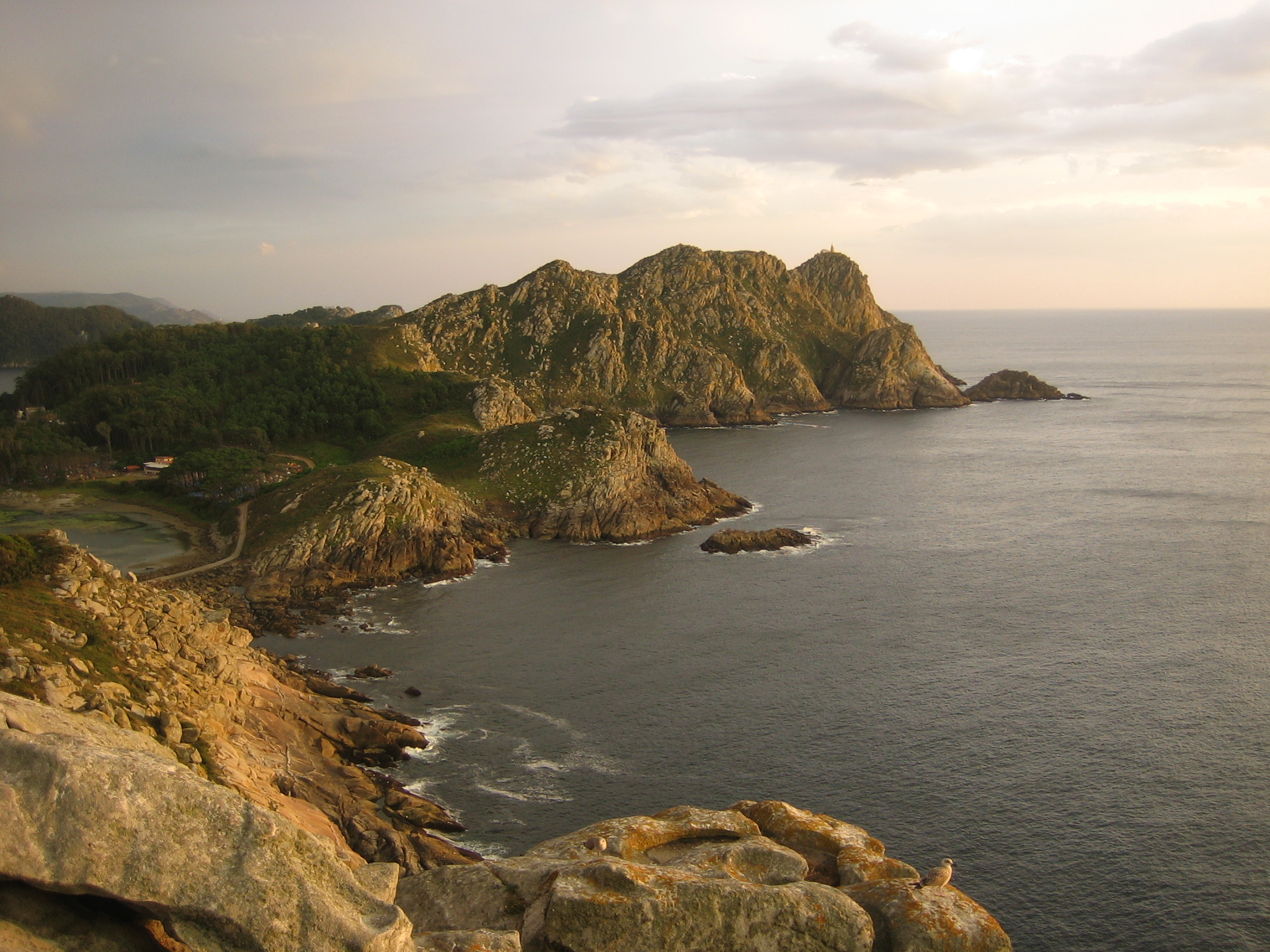 Rocky coastal cliffs and islands at sunset, with the sea extending to the horizon under a partly cloudy sky