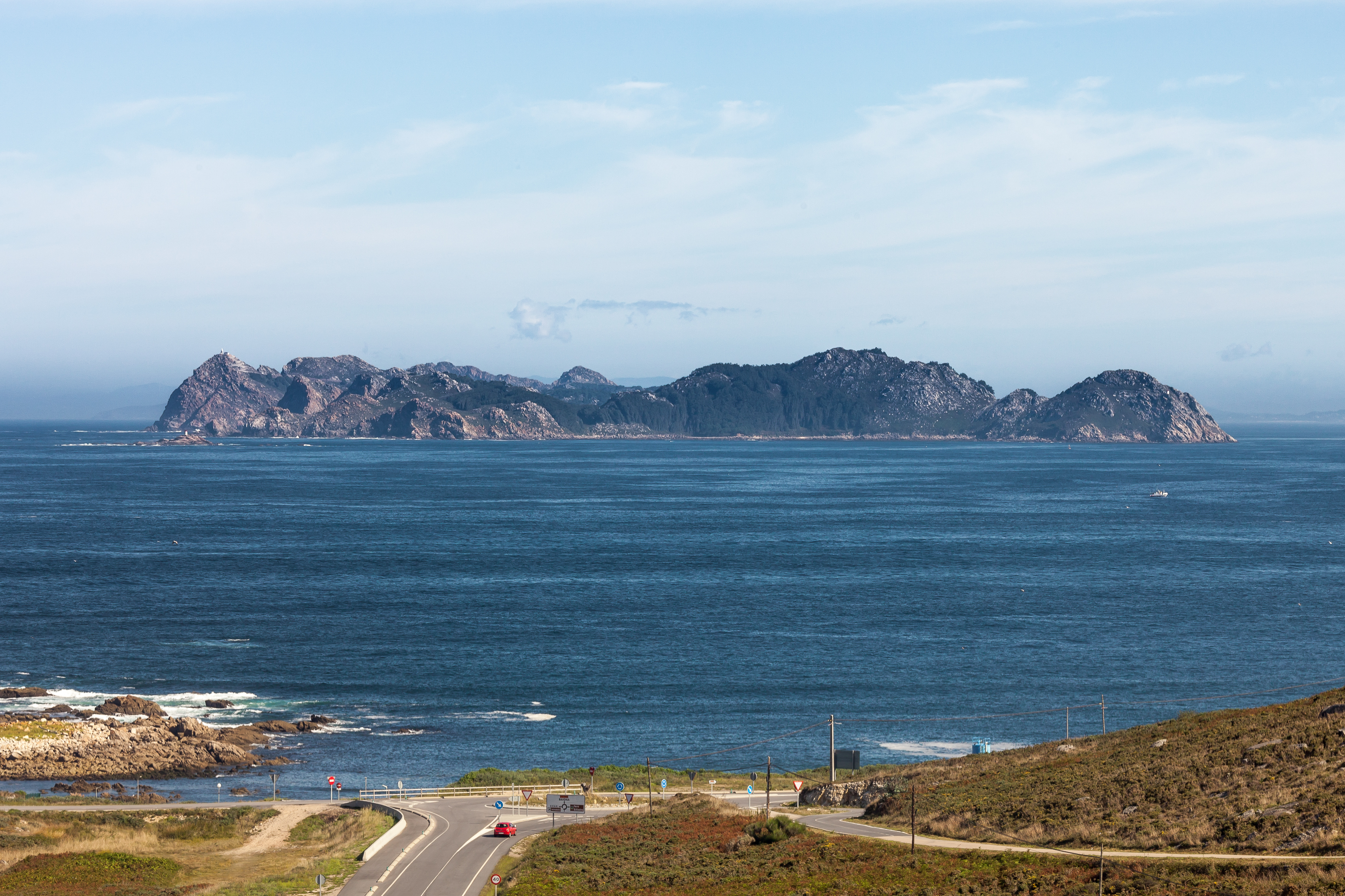 Coastal landscape showing blue ocean, distant rocky islands, a winding road, and dry grassy terrain under a partly cloudy sky
