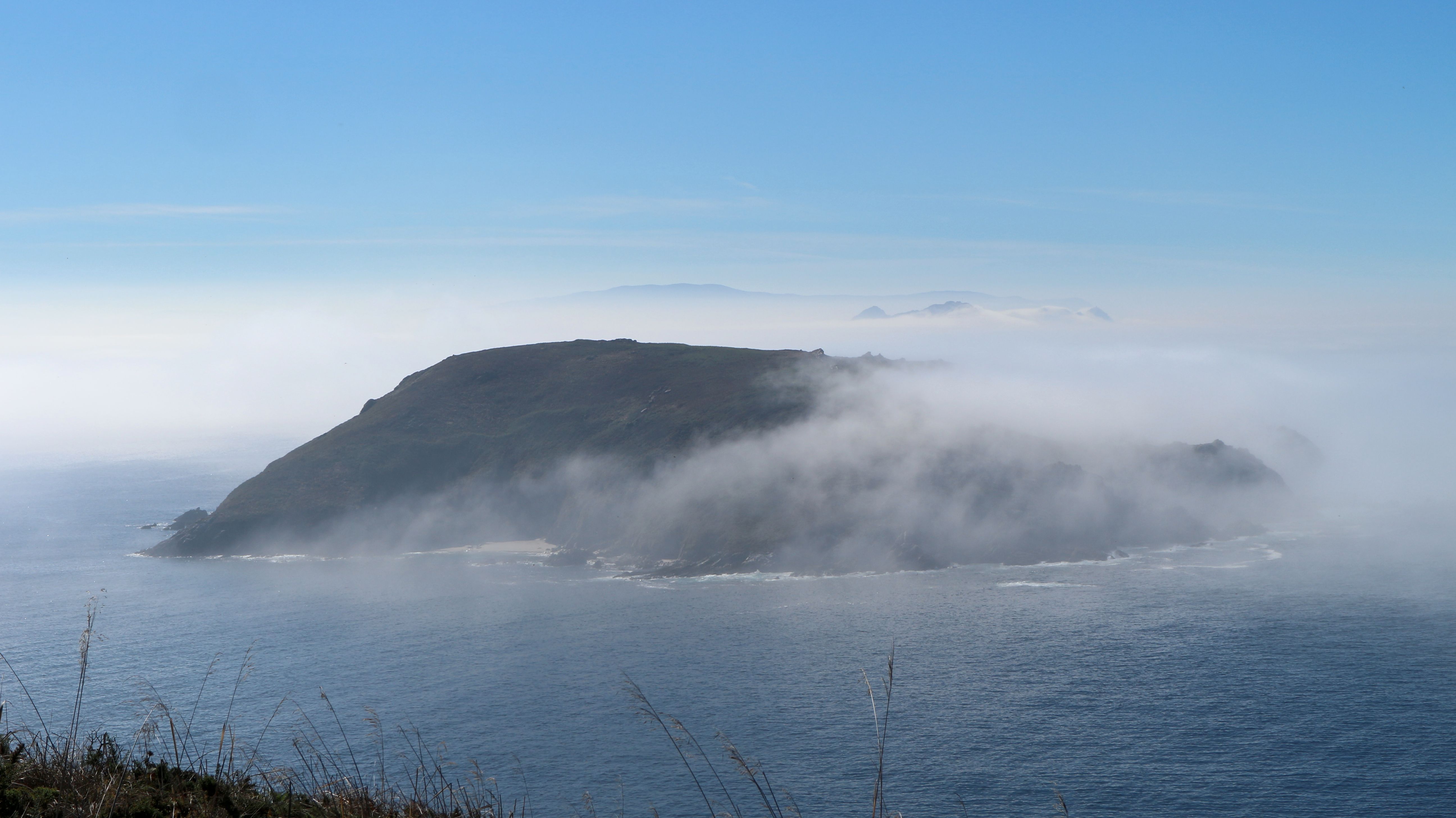 Coastal landscape showing Ons Island and Onza Island partially obscured by fog, with the sea in the foreground and a clear blue sky