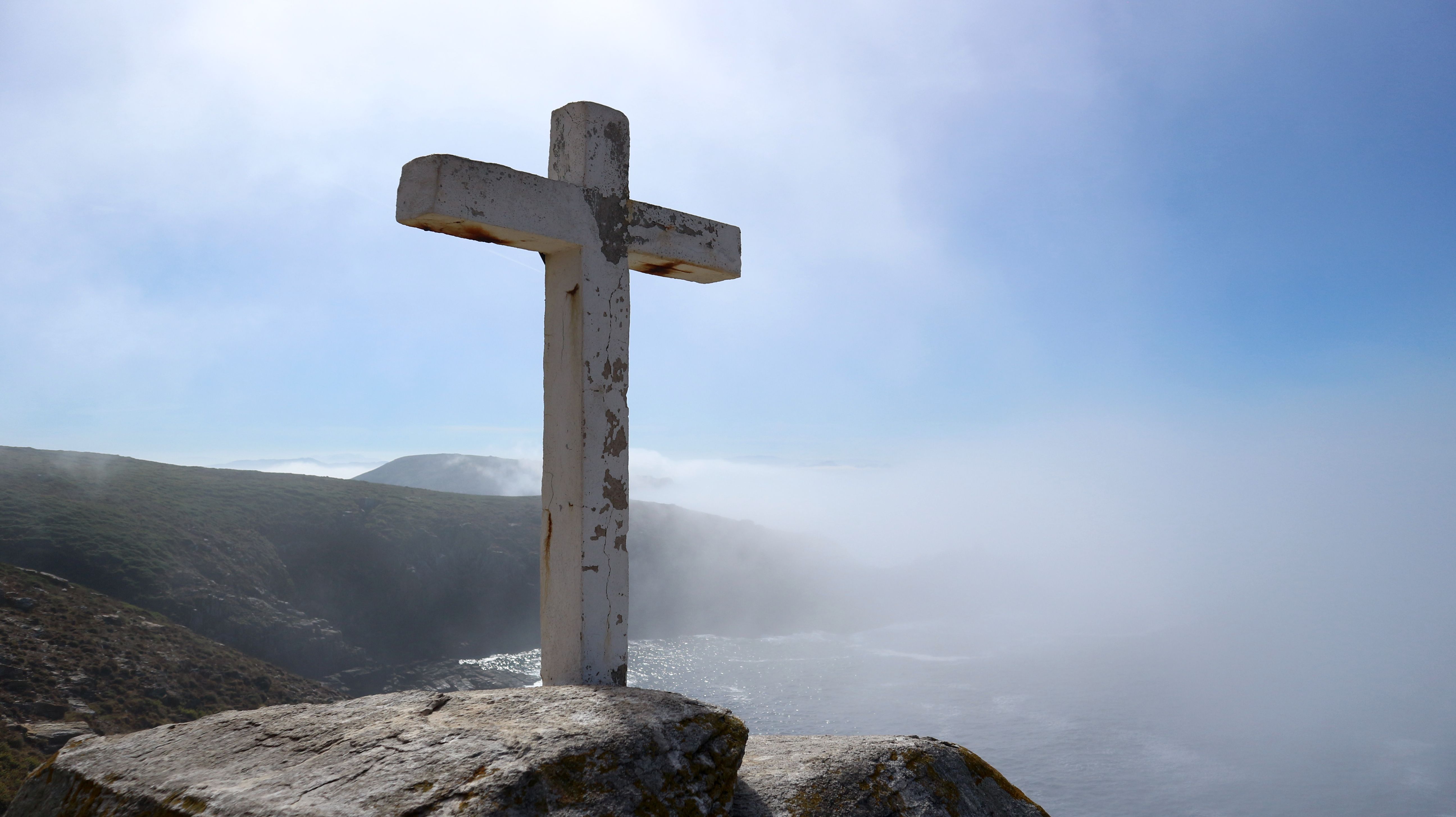 White cross on rocky terrain with misty sea and hills in background