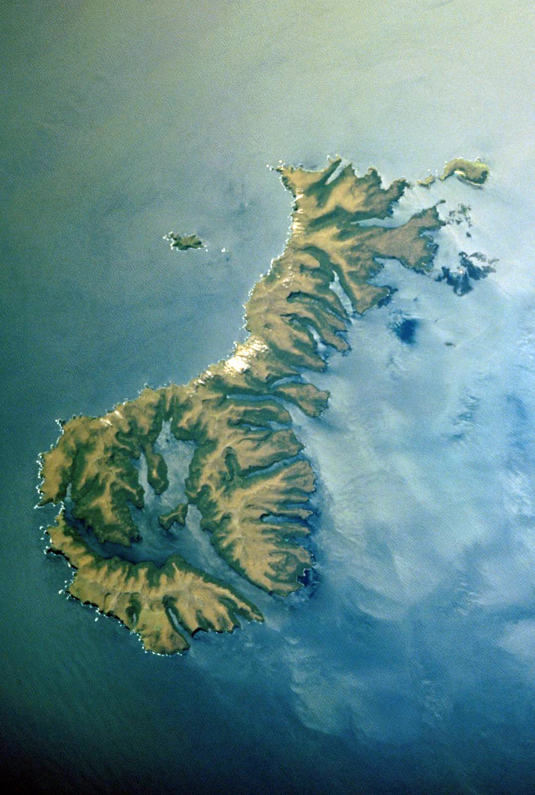 An overhead aerial view of the Auckland Islands in the South Pacific, featuring a rugged, indented coastline and steep ridges surrounded by blue ocean water.