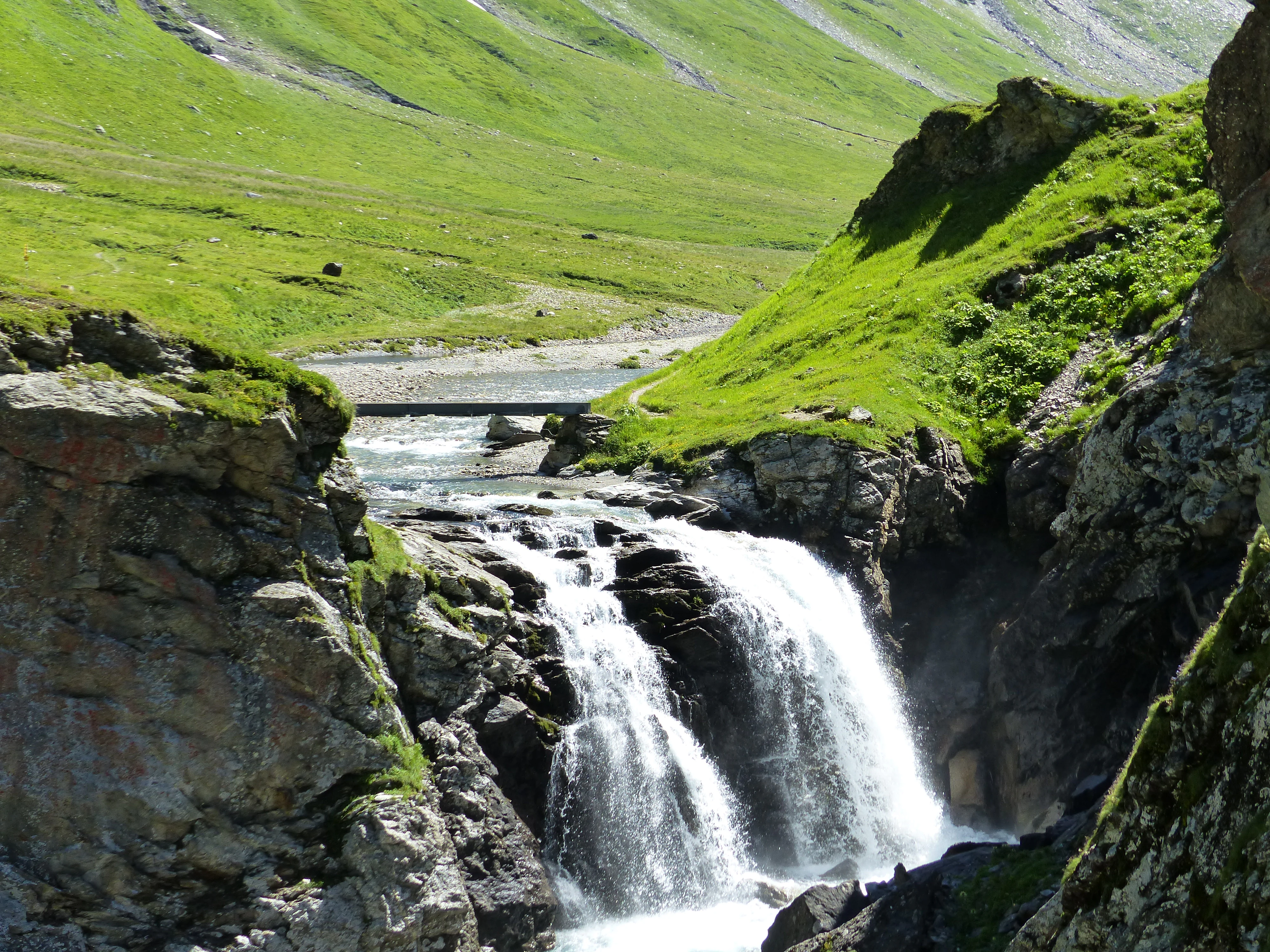 A rushing alpine waterfall cascades between rocky cliffs, with a small footbridge spanning the river in the background of a lush green valley.