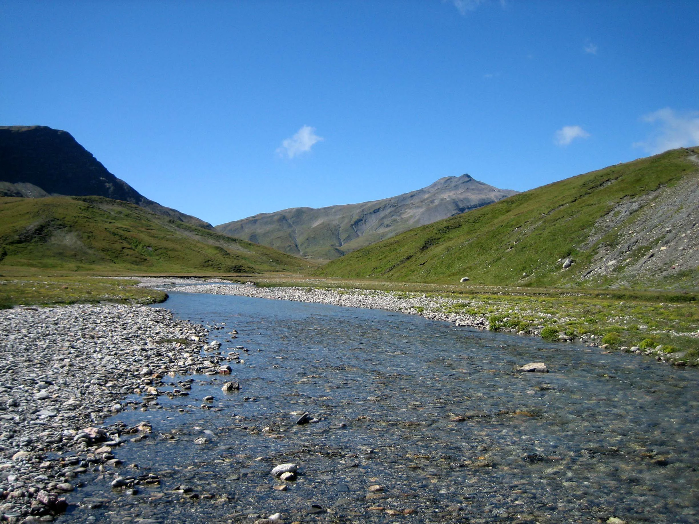 A wide shot of a shallow alpine river flowing through a green grassy valley surrounded by rolling, rocky mountains under a clear blue sky.