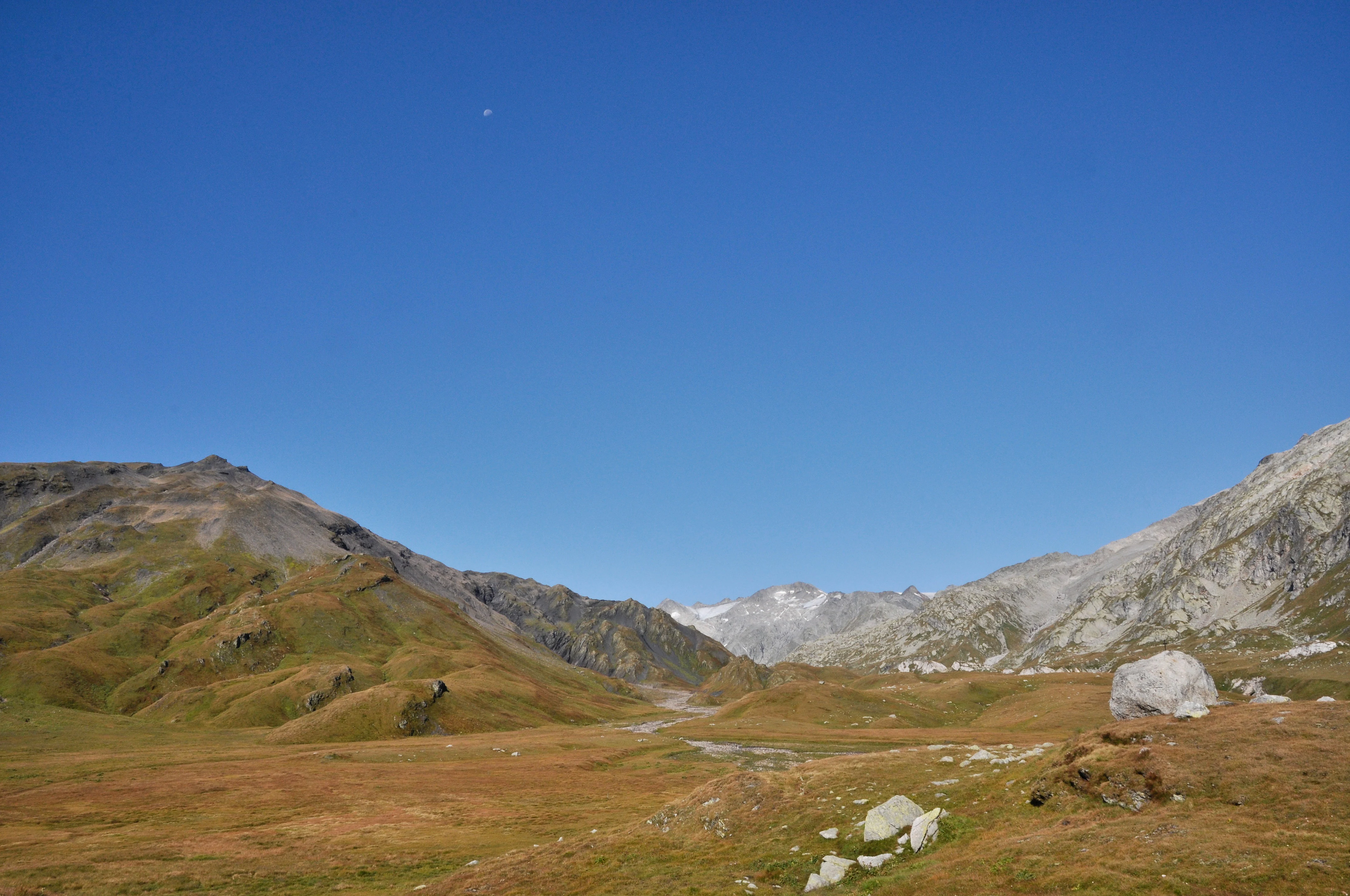 A wide, eye-level shot of a high-altitude alpine plateau with rolling, grassy hills and rocky mountain peaks under a clear blue sky. A large boulder sits in the lower right foreground.