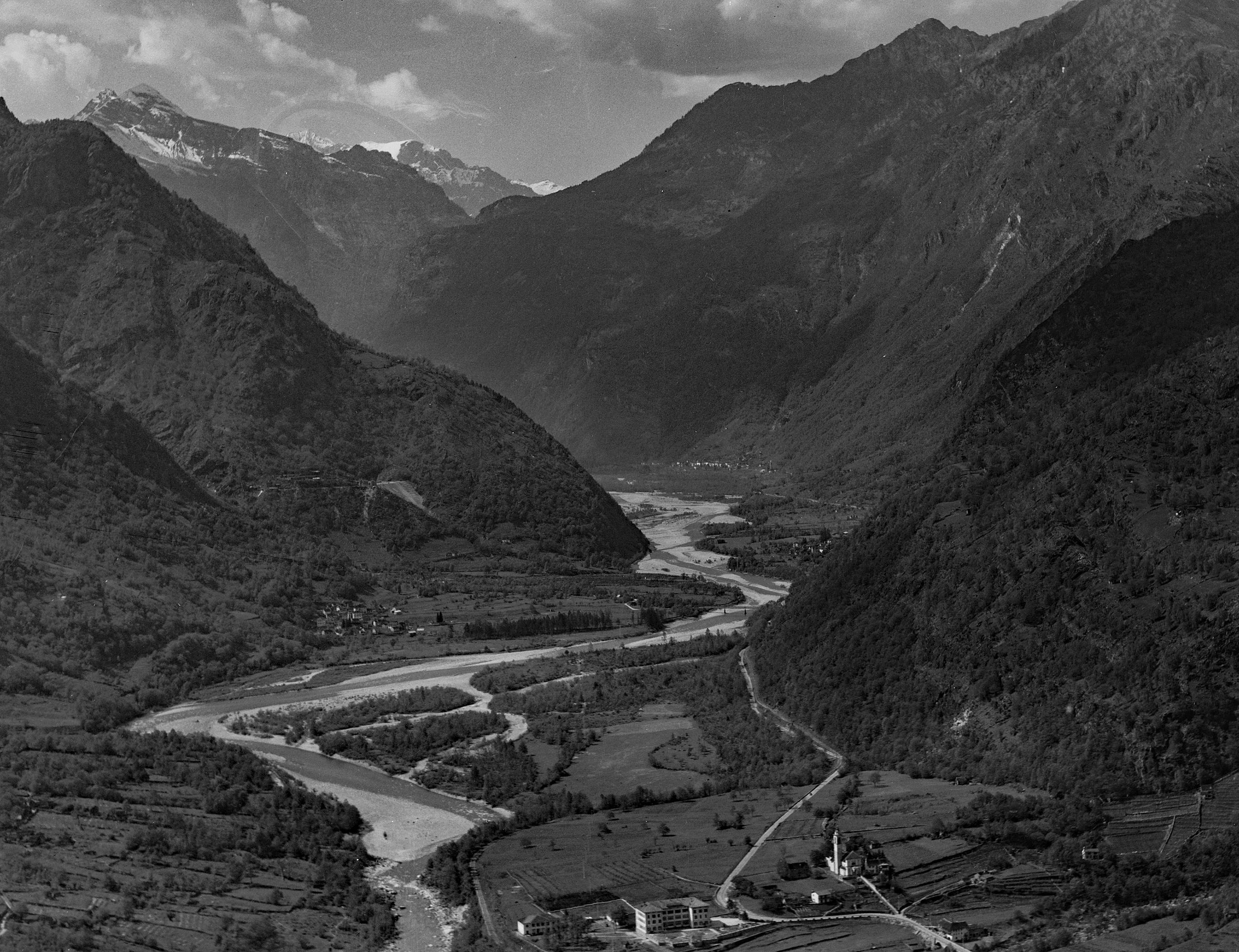 Una fotografía aérea en blanco y negro del valle de Maggia en Suiza, con un amplio río serpenteando a través de un profundo valle montañoso, con campos agrícolas y pequeñas estructuras en primer plano.
