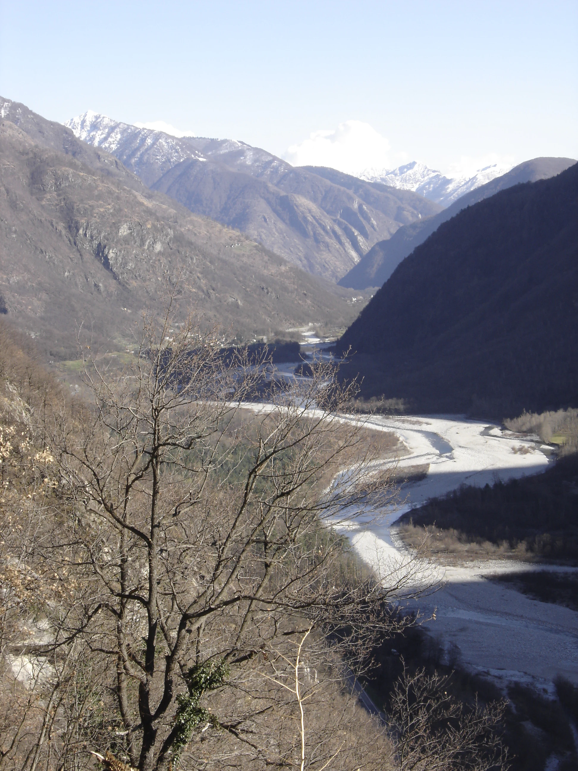 Una vista panorámica en picado hacia el valle de Maggia, con el río Maggia de lecho de grava serpenteando entre montañas escarpadas y oscuras, con picos nevados en la lejanía.