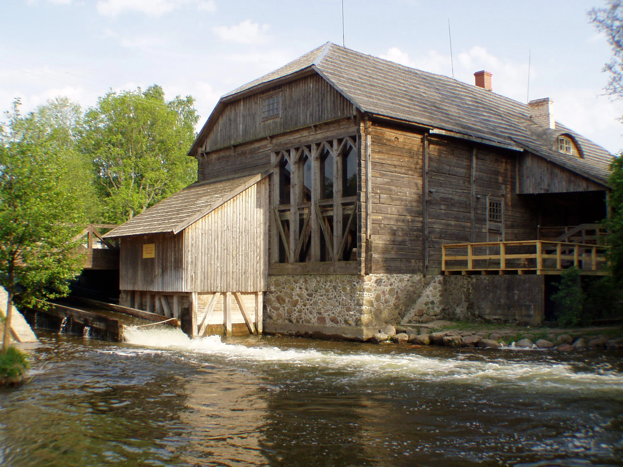 Wooden watermill building with stone foundation situated on a flowing river, surrounded by trees