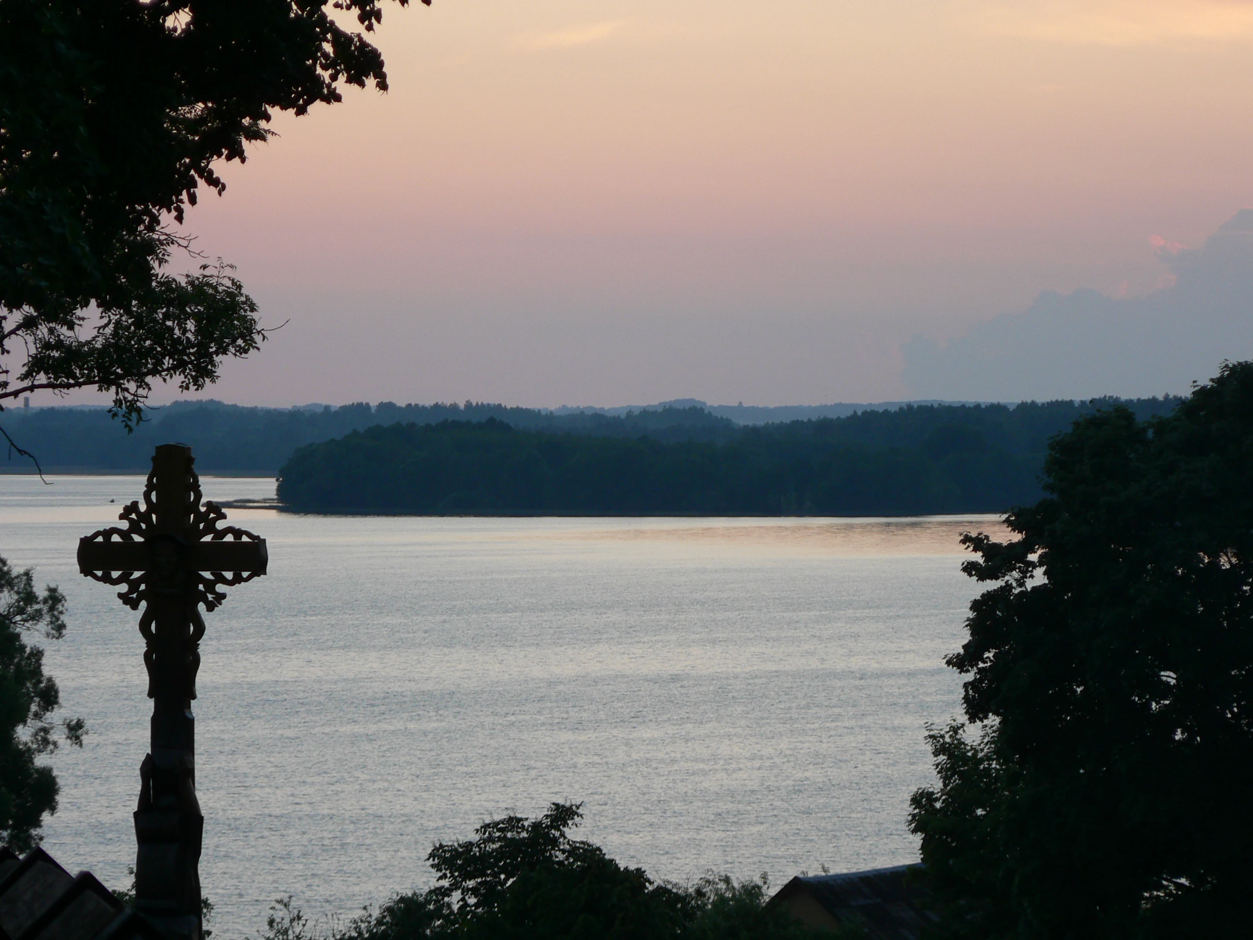 Silhouetted wooden cross in foreground with Lūšiai lake extending to the horizon under pink and blue sky with distant forested shoreline