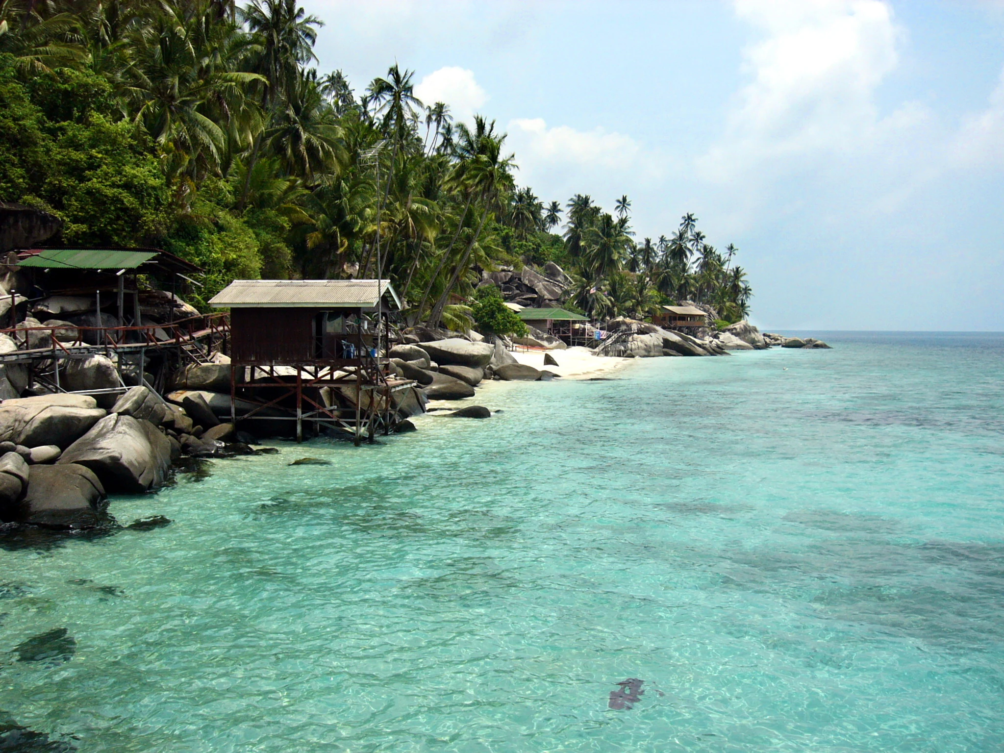 Tropical coastline on Aur Island featuring clear turquoise water, a sandy beach, large boulders, and wooden stilt huts nestled under palm trees.