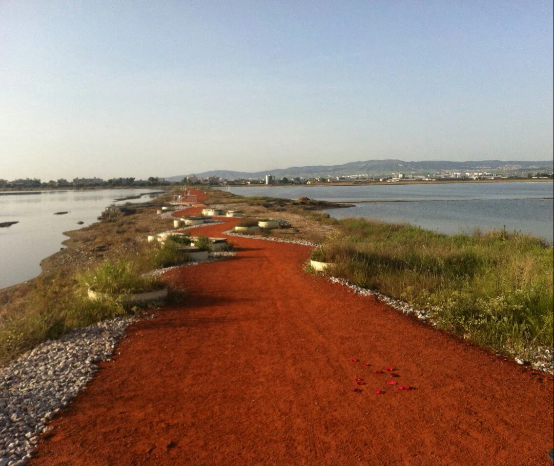 Red gravel trail running through wetland areas with water on both sides under clear blue sky