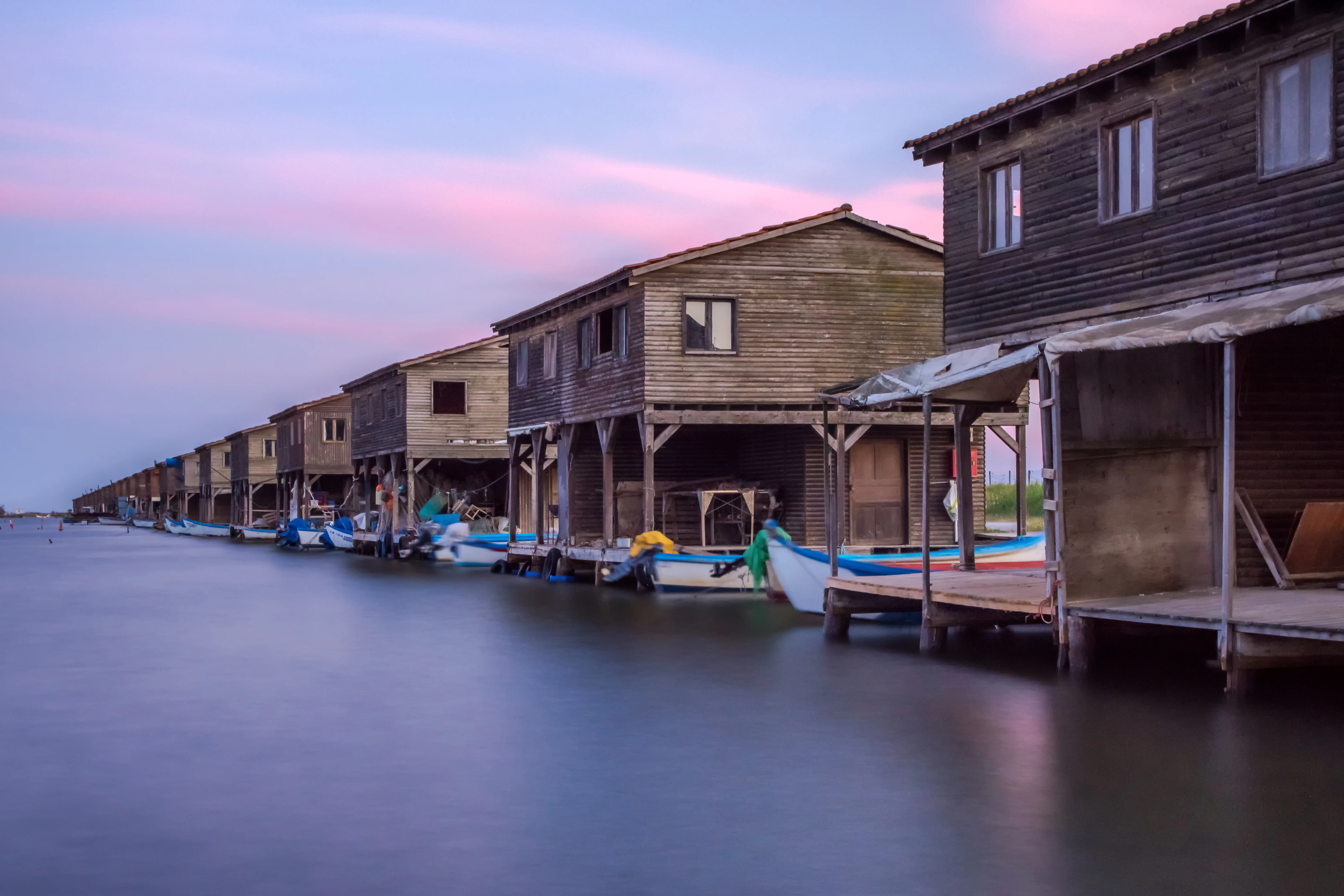 A row of wooden houses on stilts along a calm waterway with boats docked along a wooden pier