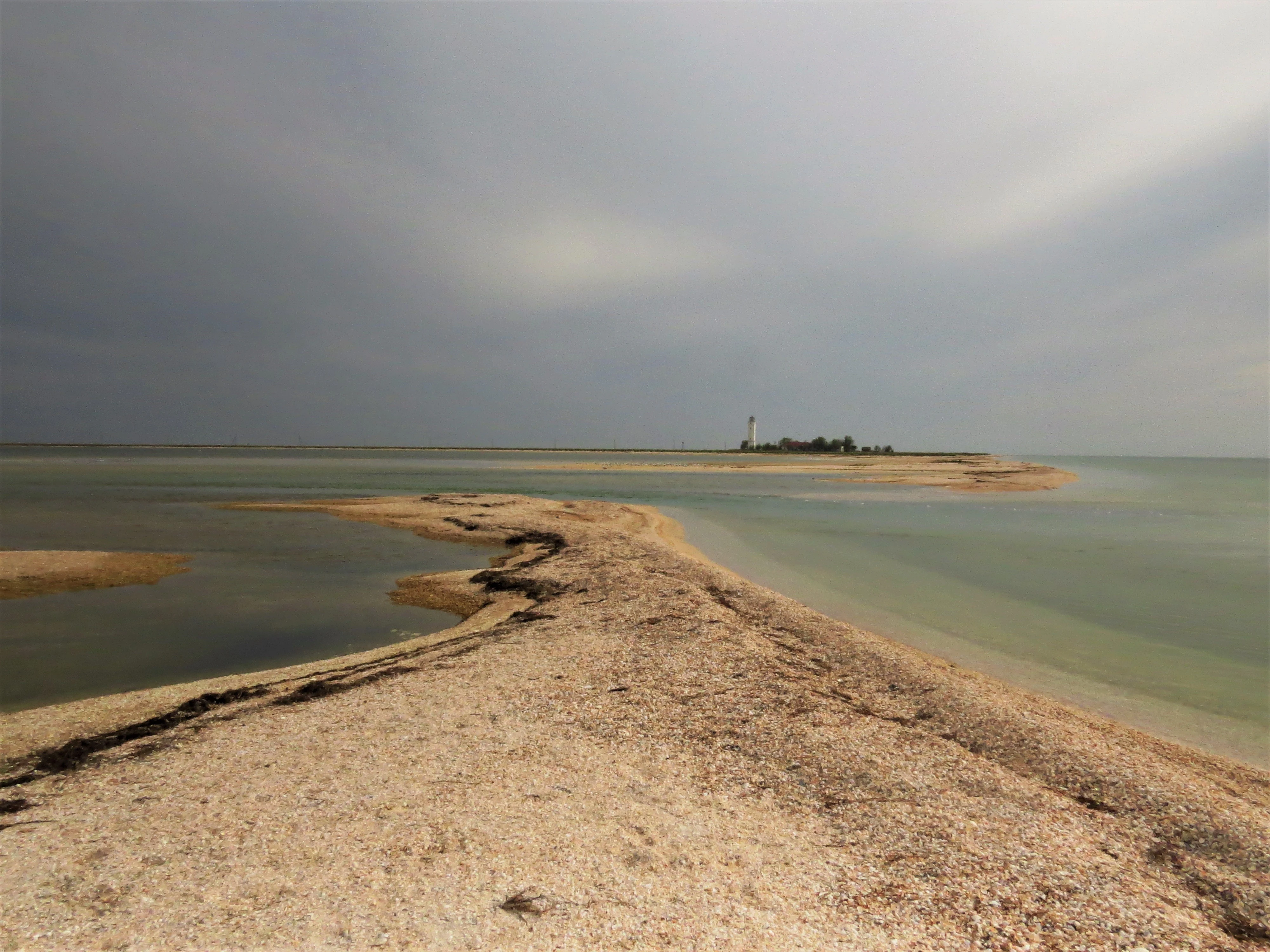 Sandy coastal spit with shallow water on both sides leading to a small island with a distant lighthouse under cloudy skies