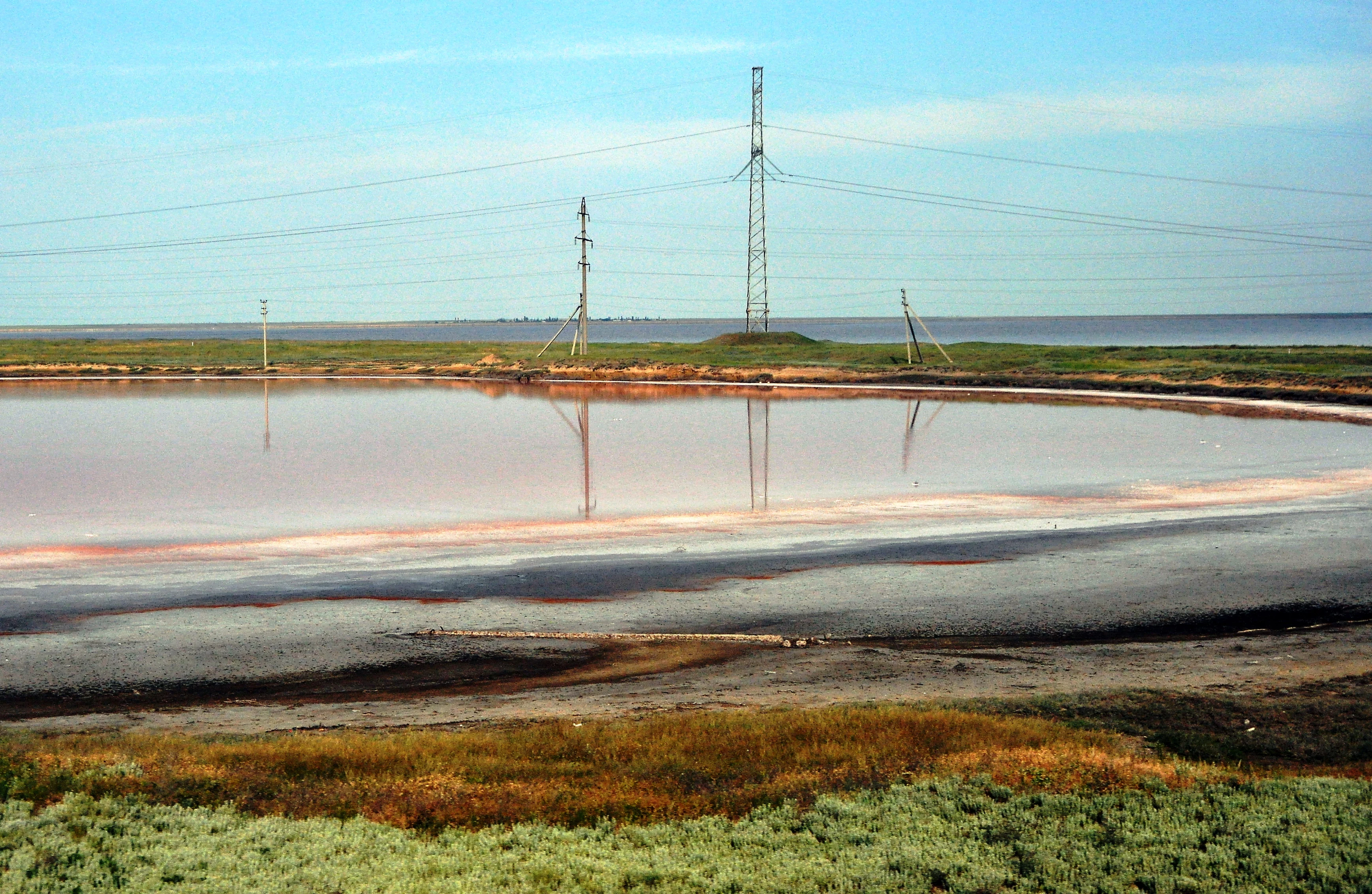 Pink-tinted wetland water body with grassy terrain, power lines, and transmission towers under a clear blue sky
