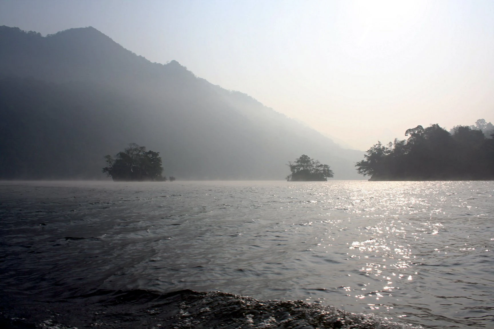 Large body of water with mist, two small forested islands, and mountains in the background under a hazy sky