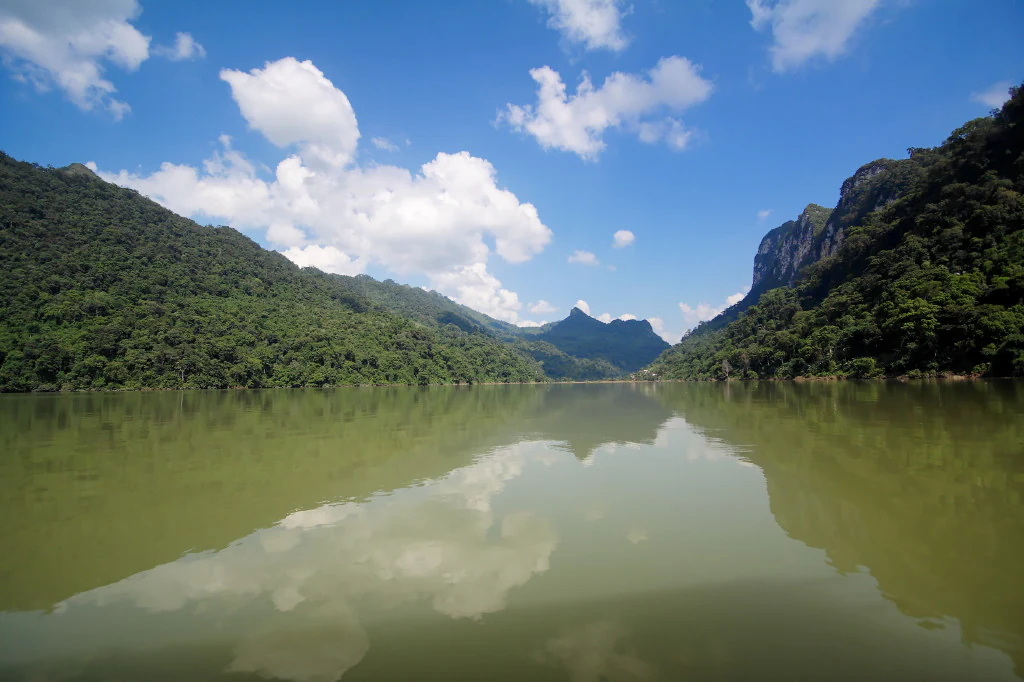 A calm lake reflecting a bright blue sky with white clouds, surrounded by lush green mountains