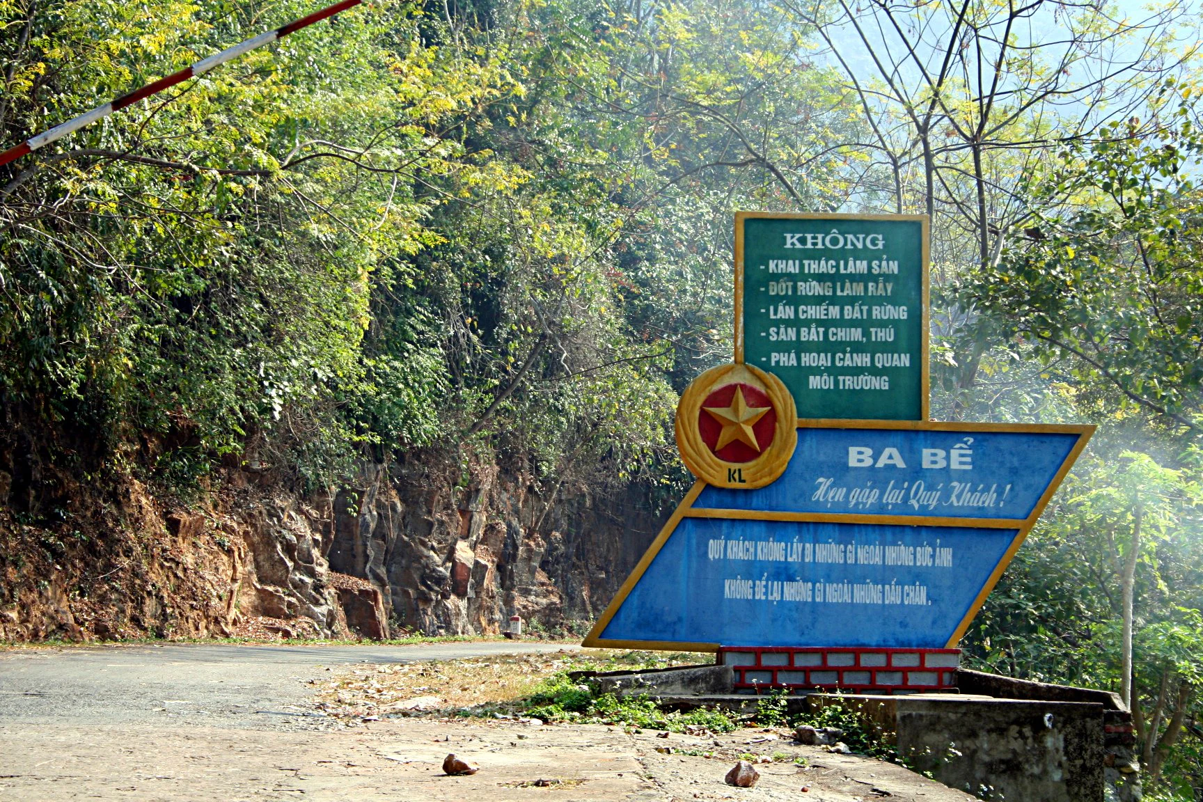 Green and blue entrance sign for Ba Bể National Park with Vietnamese text, red star emblem, and barrier arm, surrounded by forest and road