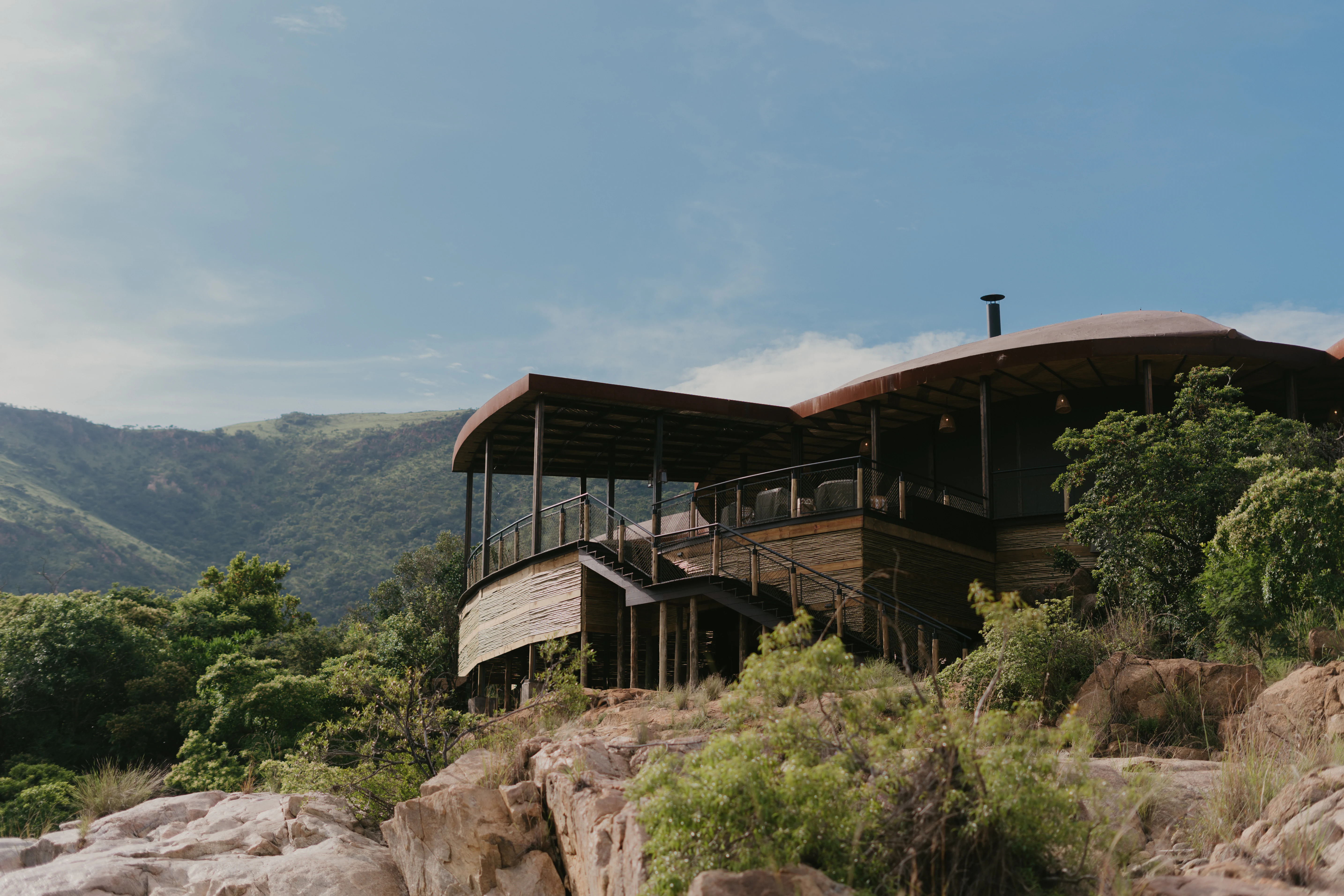 A modern lodge structure with wooden siding and a curved roof sits on a rocky ridge overlooking a verdant valley in Babanango Game Reserve.