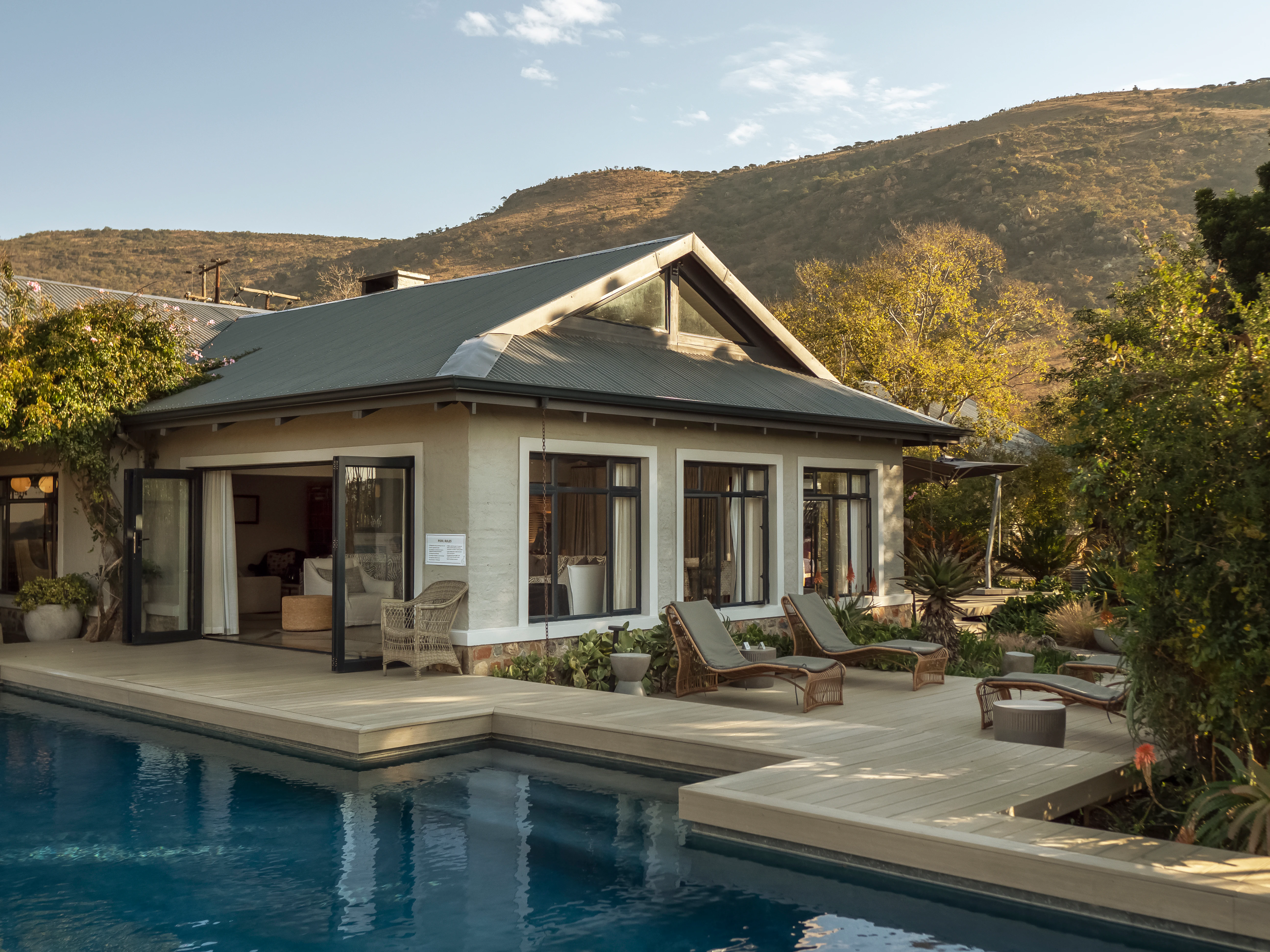 A modern, single-story lodge building with large windows and open doors, featuring an adjacent swimming pool and lounge chairs on a wooden deck, with a large hill in the background.