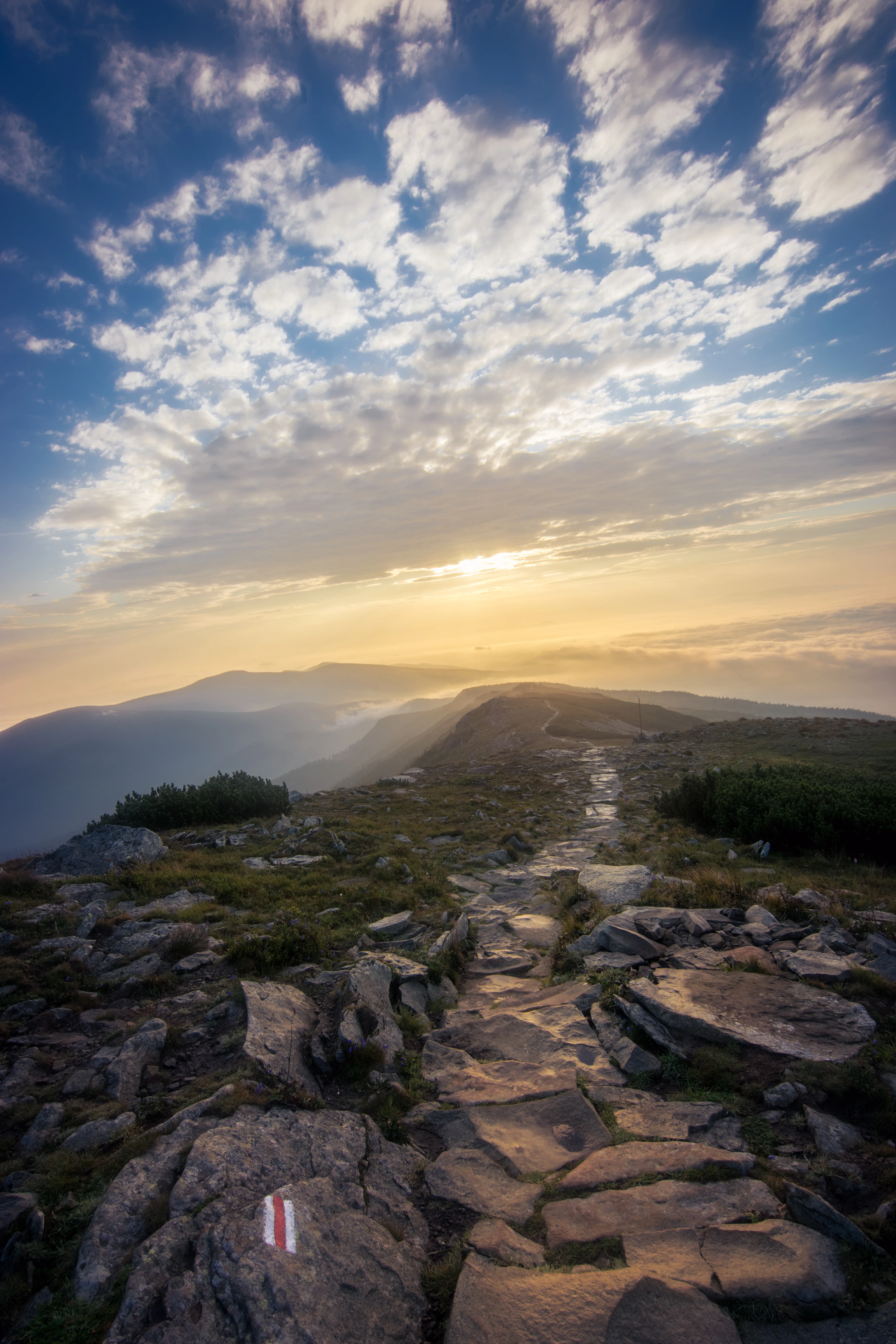 Stone trail with red and white trail marker on rocky terrain, mountain landscape with distant peaks and sunset sky