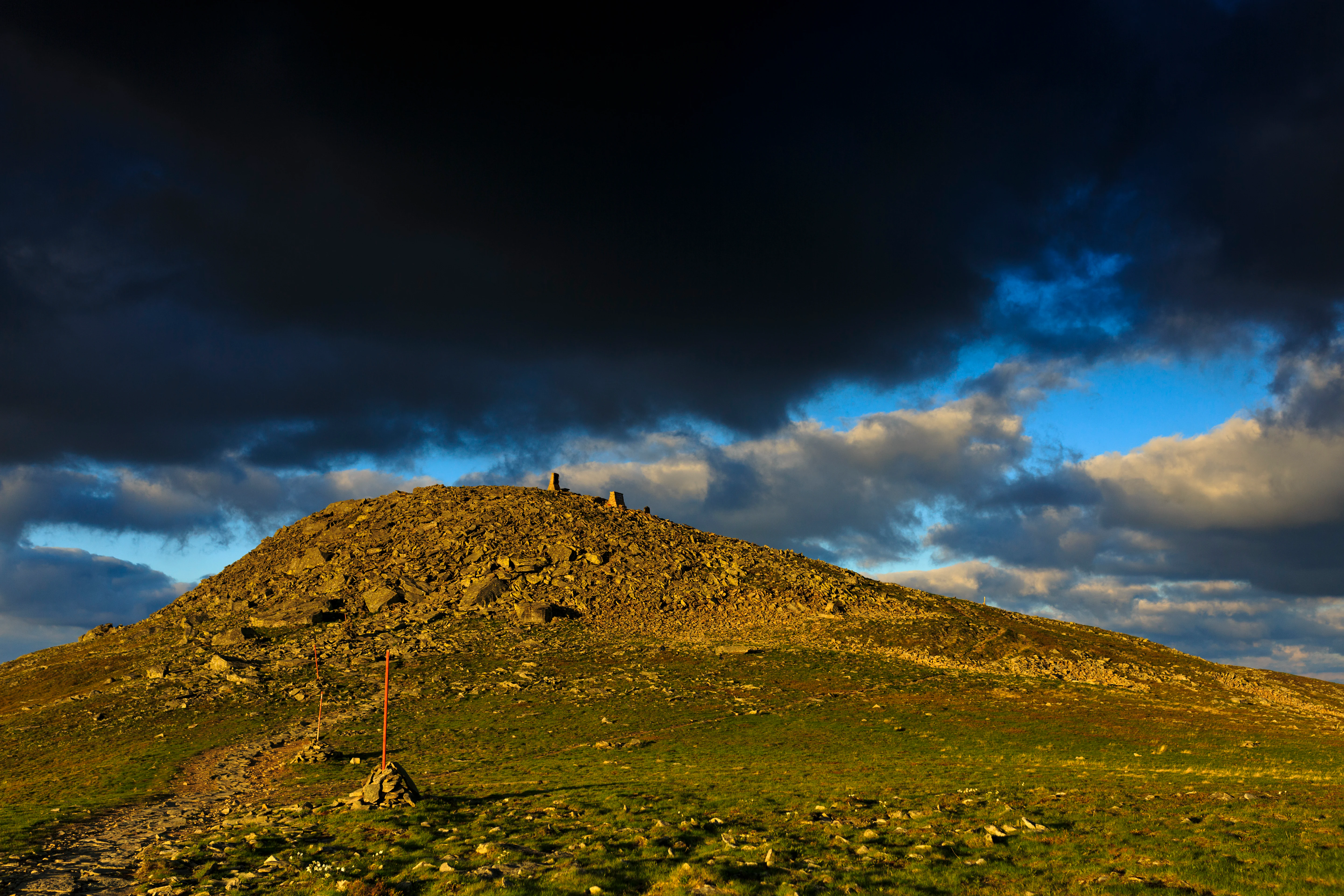 Rocky mountain peak with grassy slope and trail leading to summit under dark clouds with patches of blue sky