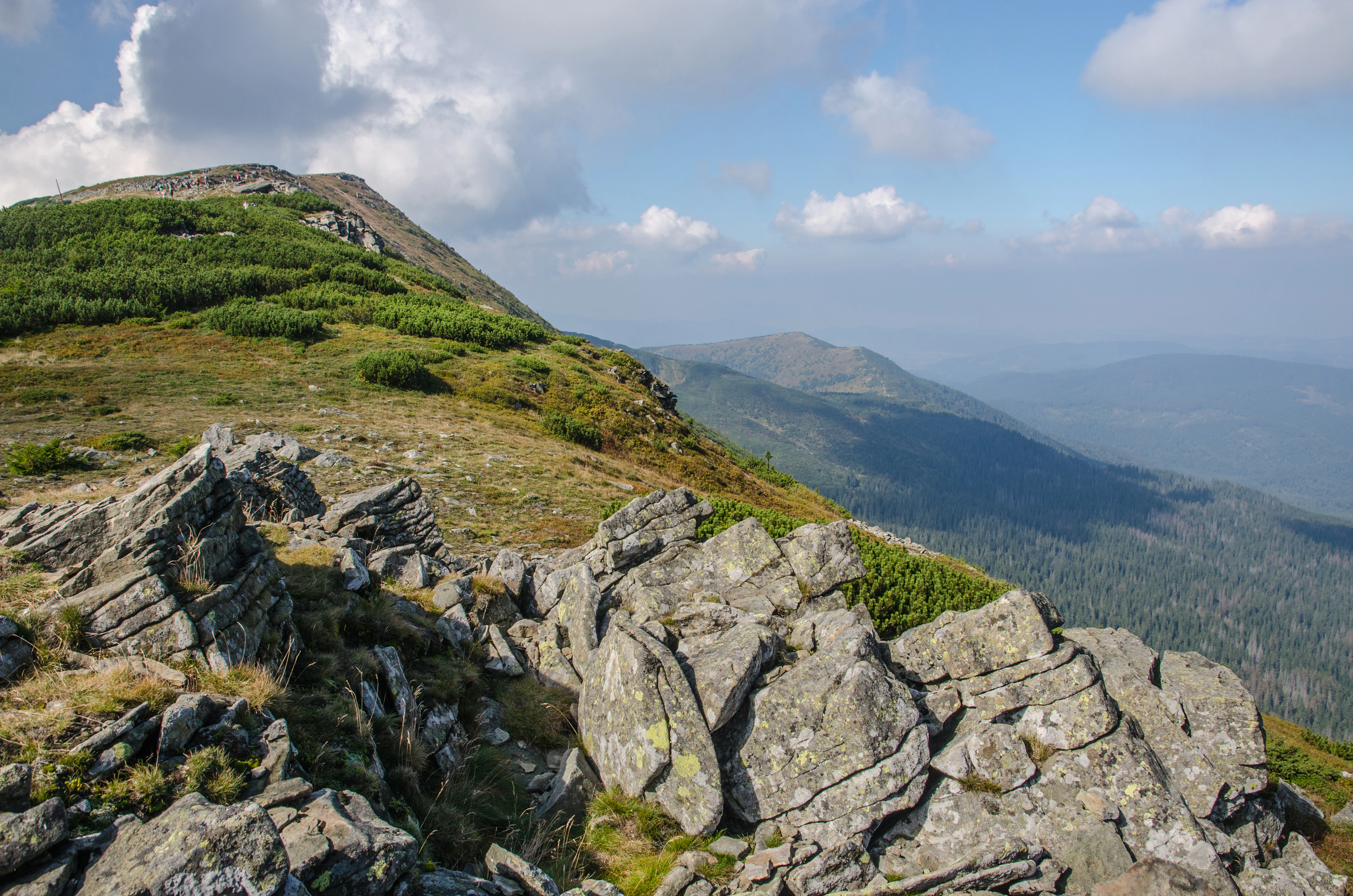 A landscape view of a rocky mountain ridge with low vegetation and distant forested slopes under a partly cloudy sky.
