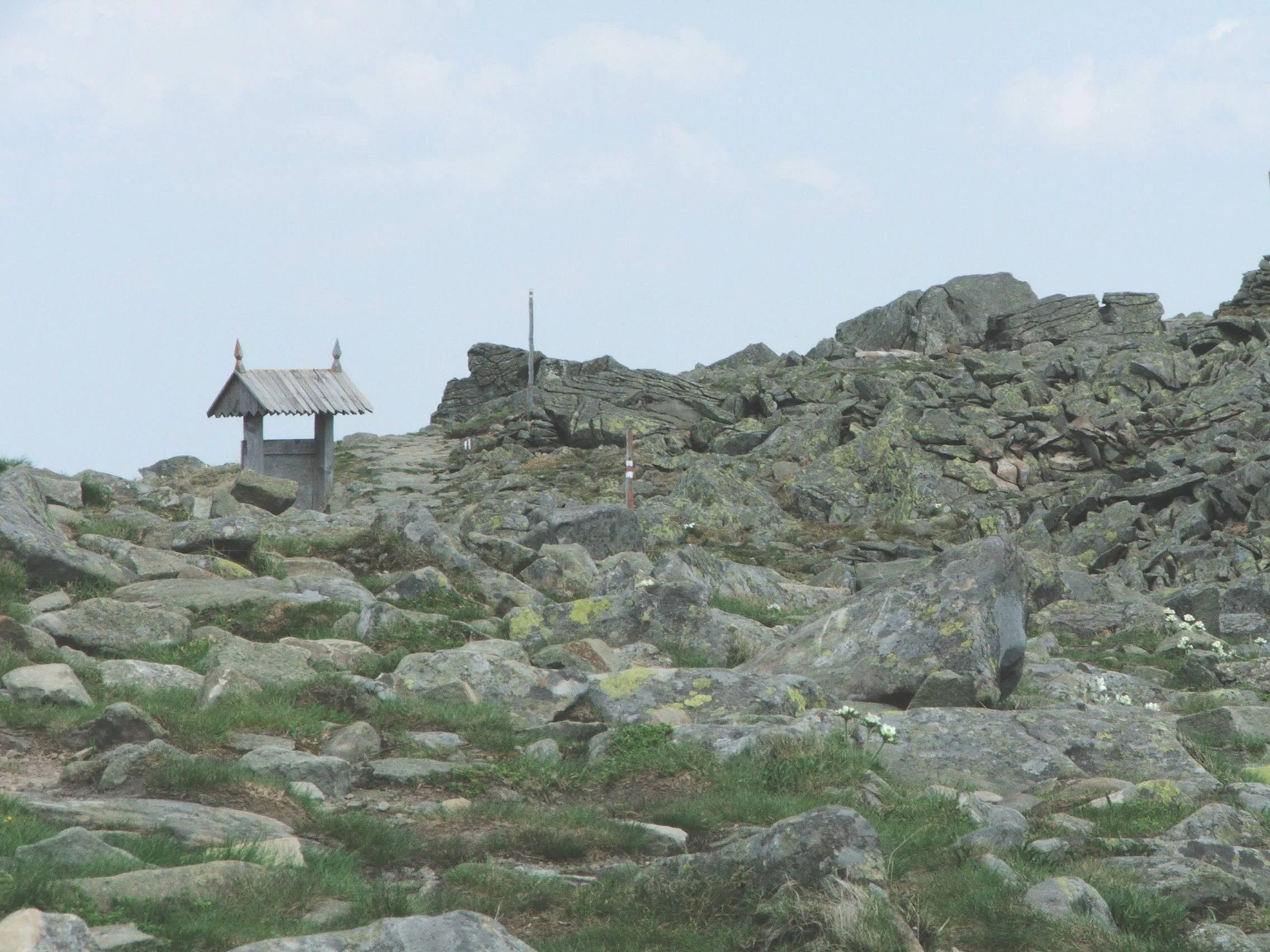 A rocky, uneven mountain slope covered in grey stones and patches of low vegetation, with a small wooden structure with a gabled roof visible in the distance.