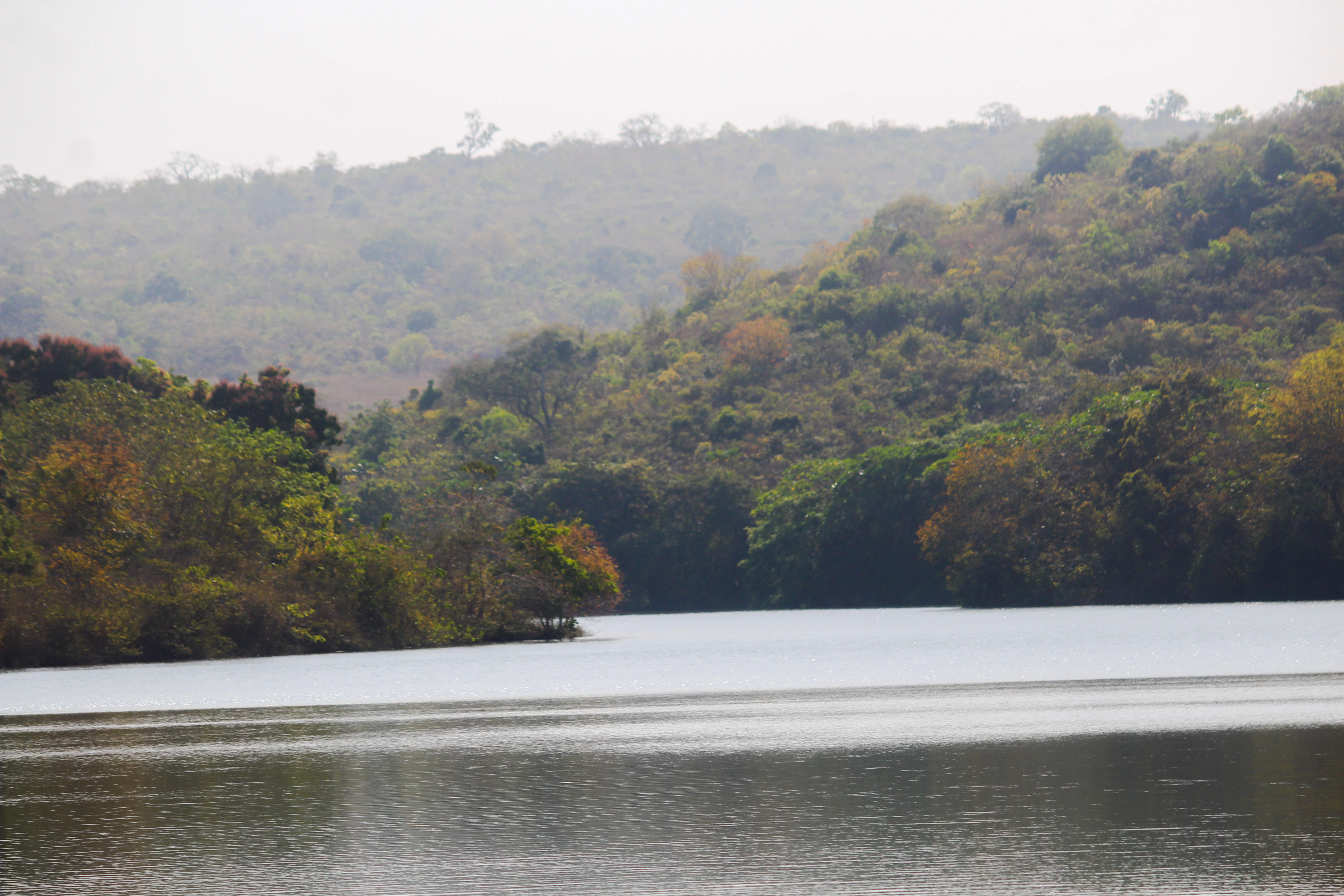 A wide shot of a calm river or lake with forested hills in the background under a hazy sky.