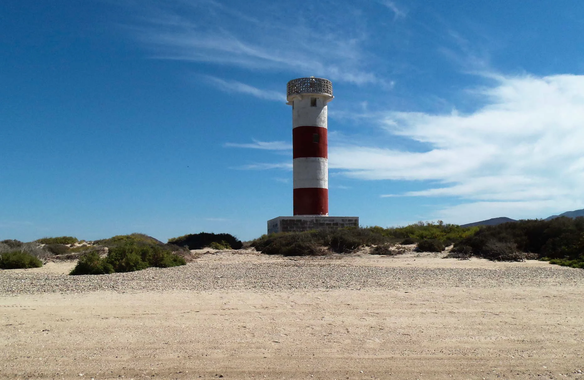 Un faro cilíndrico de rayas rojas y blancas se alza sobre un cabo costero arenoso y cubierto de matorrales, bajo un cielo azul brillante.