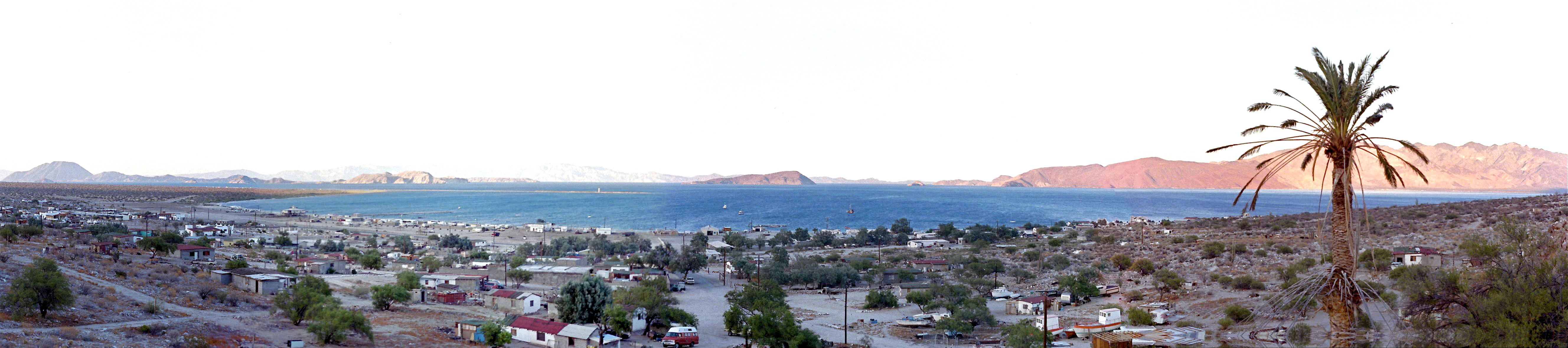 Vista panorámica de un pueblo costero enclavado en un valle desértico que conduce a una bahía con islas lejanas bajo un cielo brillante.