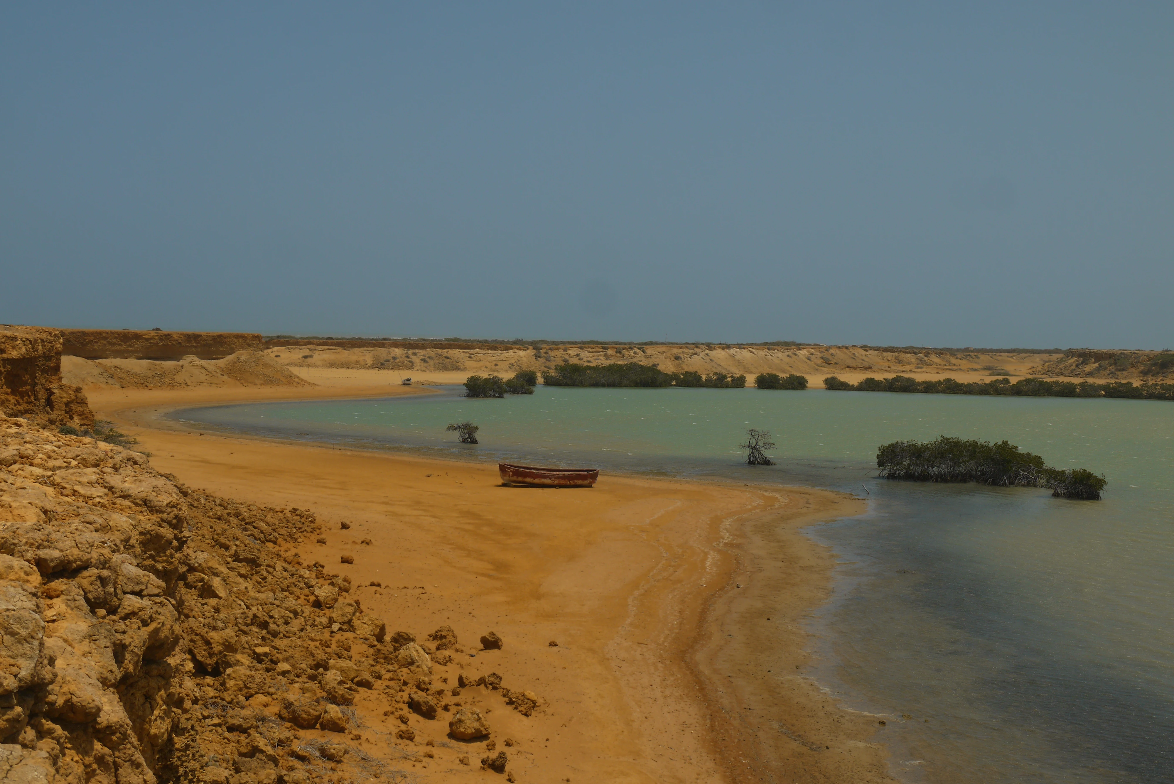 Sandy beach with shallow turquoise water, rocky cliffs on the left, and small islands in the distance under a clear blue sky
