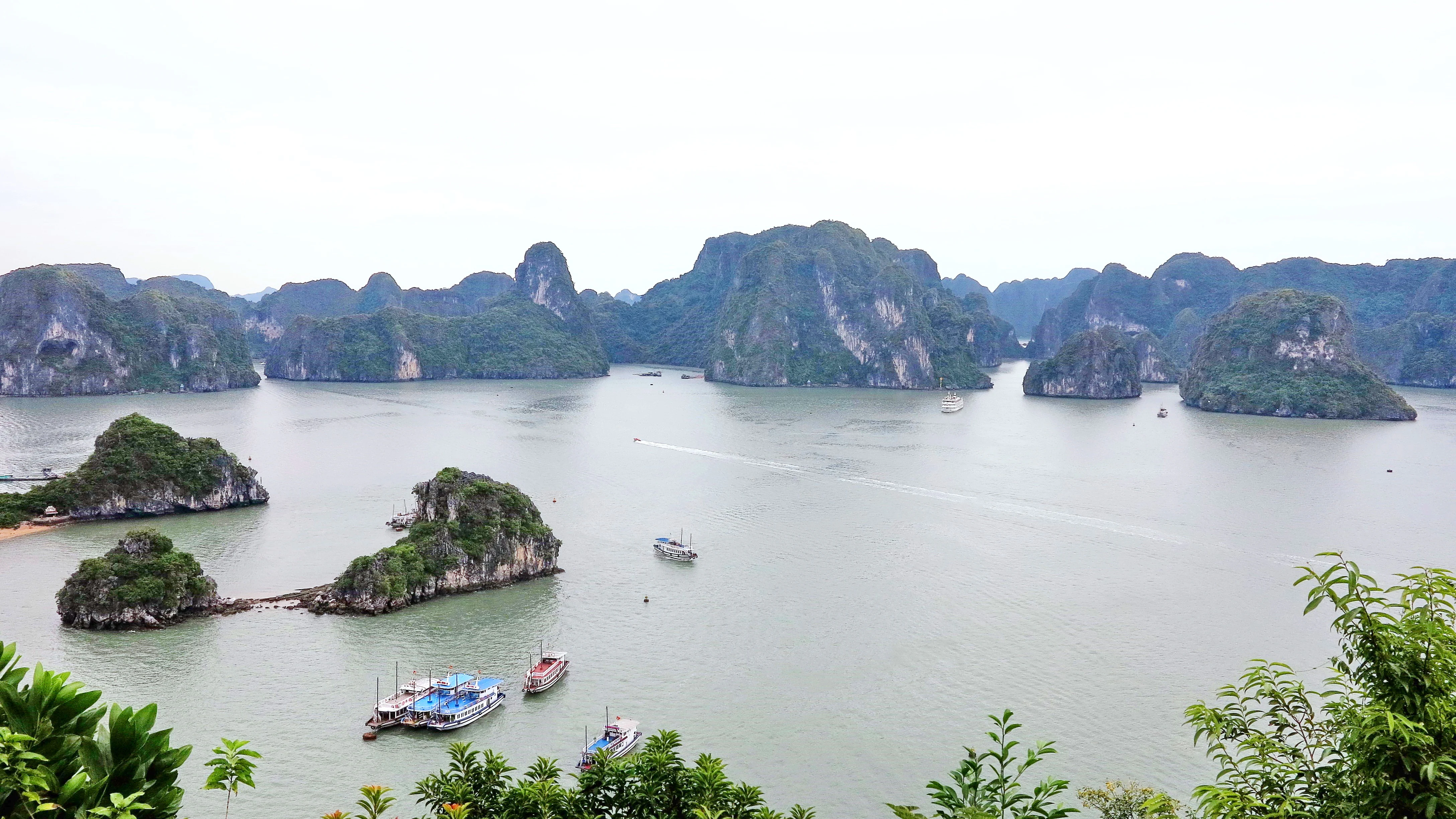 Panoramic view of Bai Tu Long Bay with limestone karst islands, calm water, boats, and green vegetation