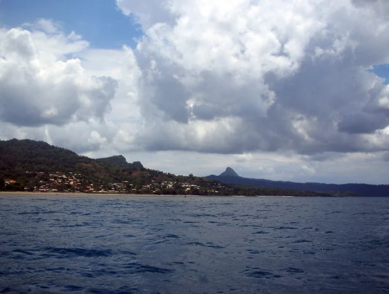 A wide shot looking across the water toward the hilly, green coastline of Baie de Bouéni in Mayotte, with a prominent peak in the distance under a partly cloudy sky.