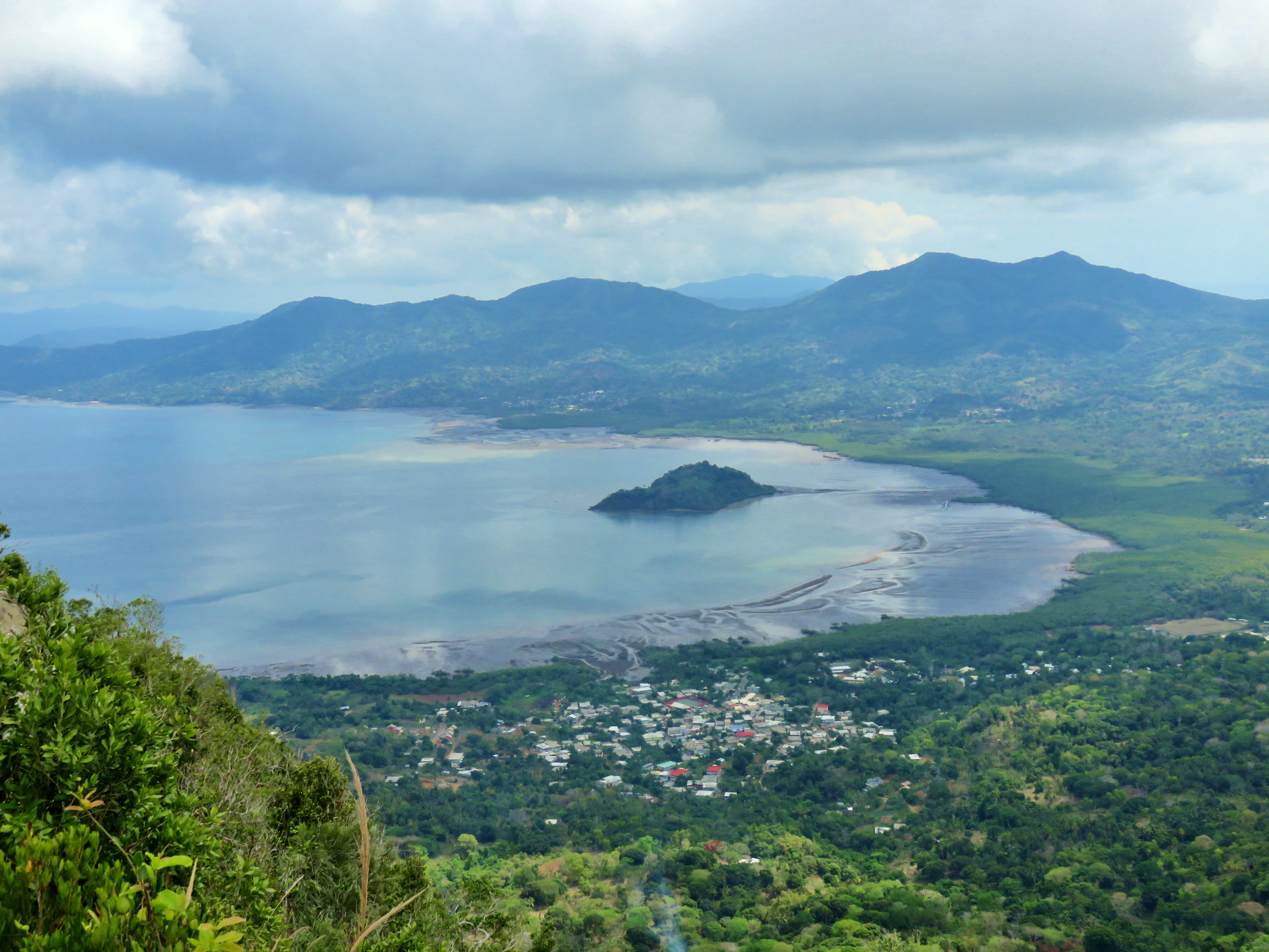 A high-angle, wide-angle view overlooking a broad, calm bay with a small, forested islet in the center and a small town nestled in the lush coastal landscape below.