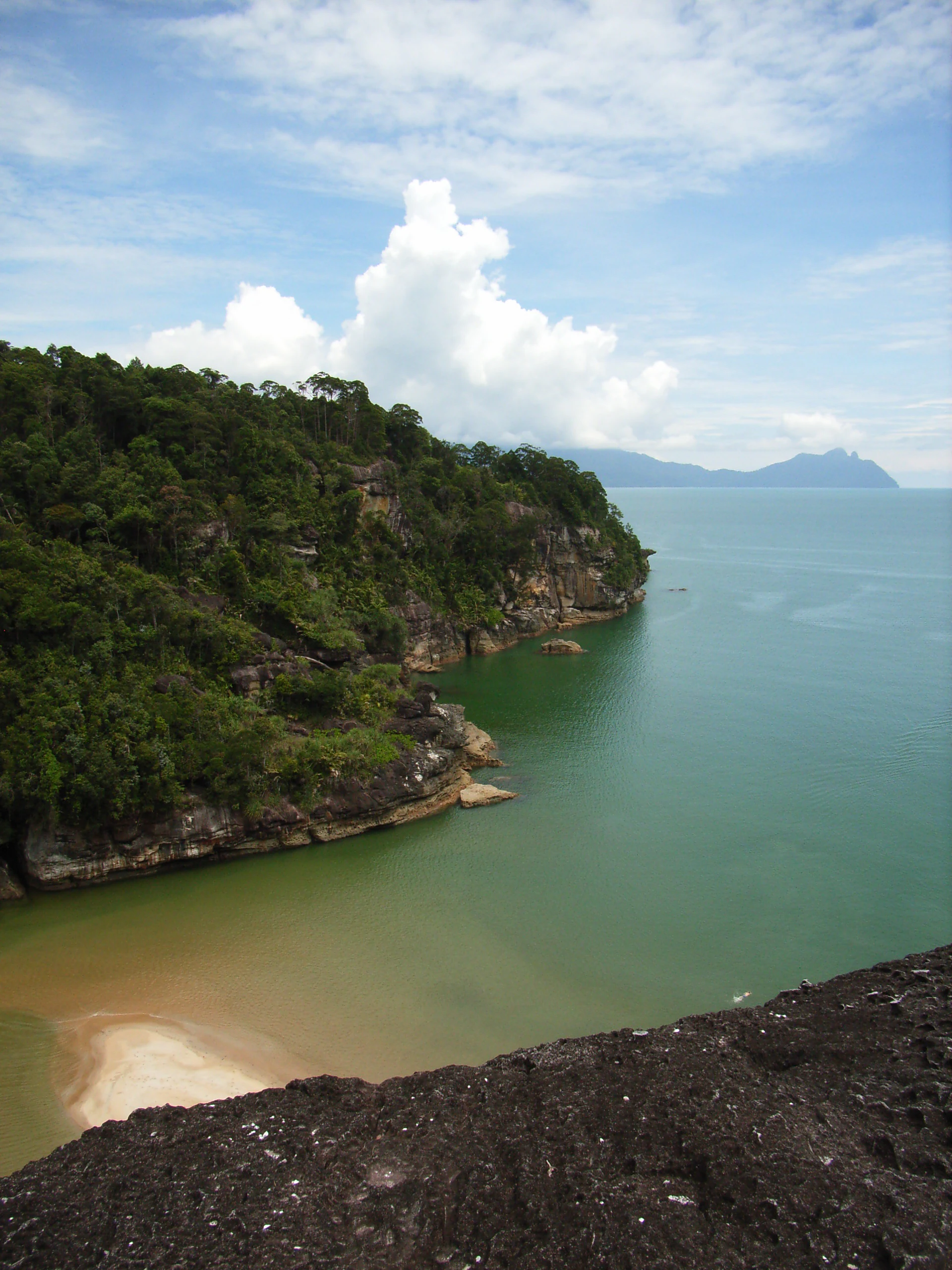 Forest-covered cliffs overlooking turquoise water and sandy beach, with distant mountains under partly cloudy sky