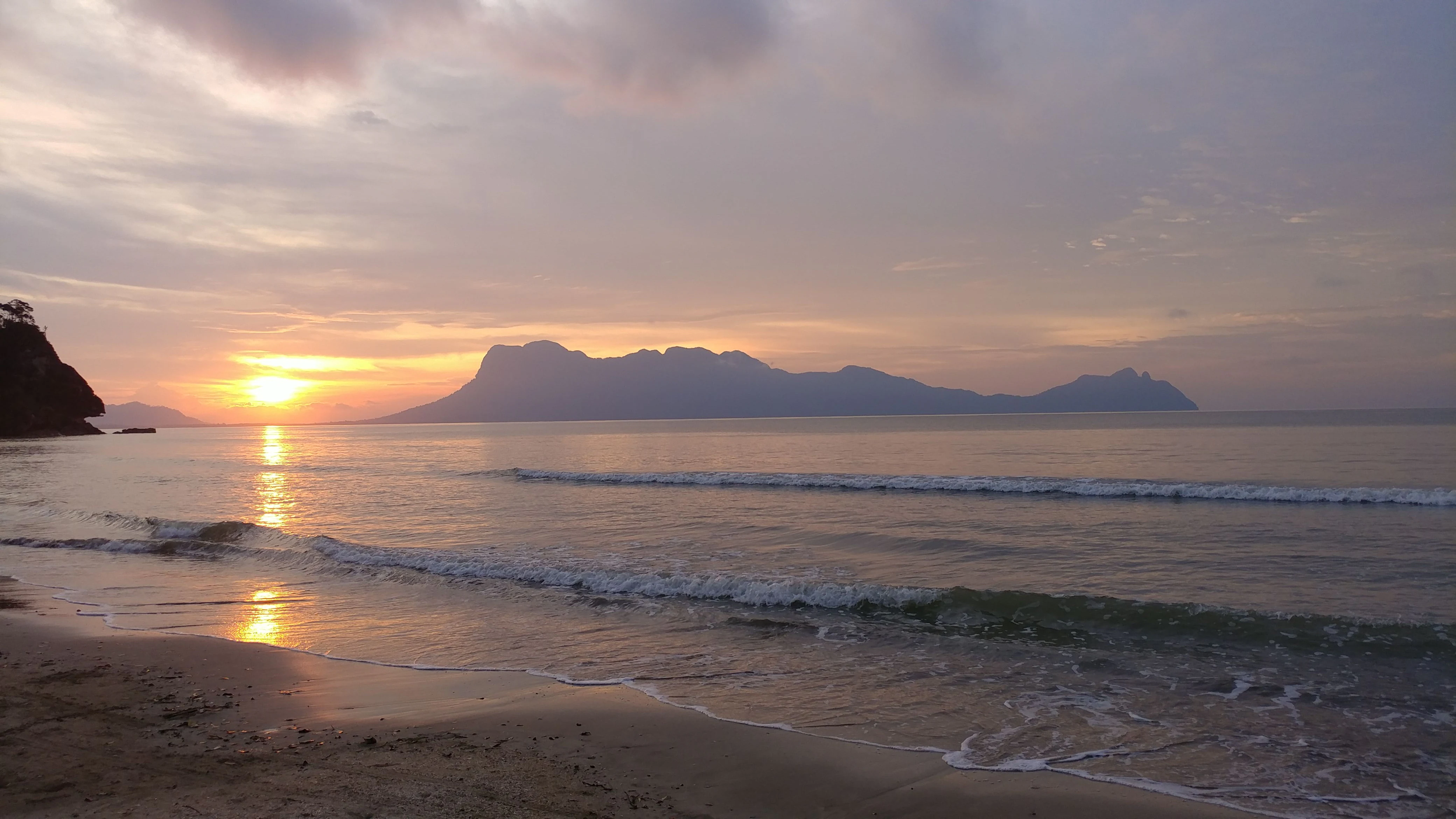 Sunset over a sandy beach with gentle waves, distant mountains, and a partly cloudy sky