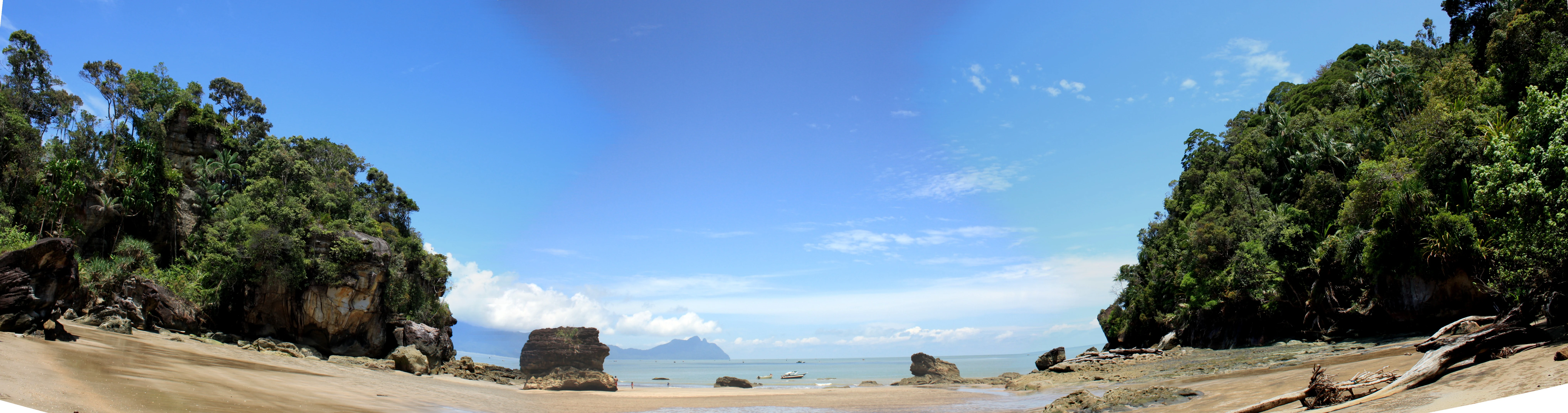 Panoramic view of a sandy beach bordered by rocky outcrops and lush green forest, with a clear blue sky overhead
