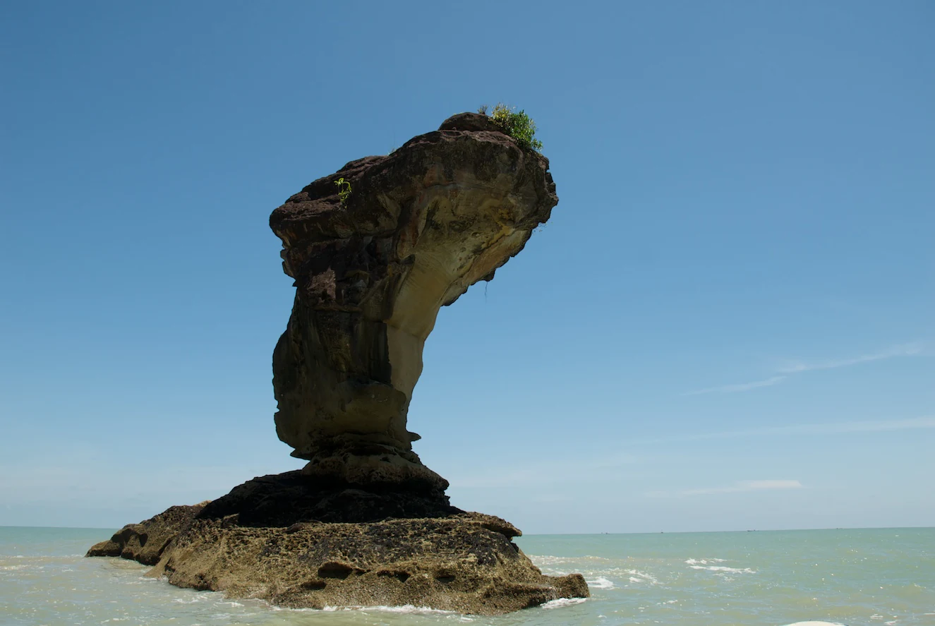 A large rock formation with a narrow base and wider top standing in shallow ocean water under a clear blue sky
