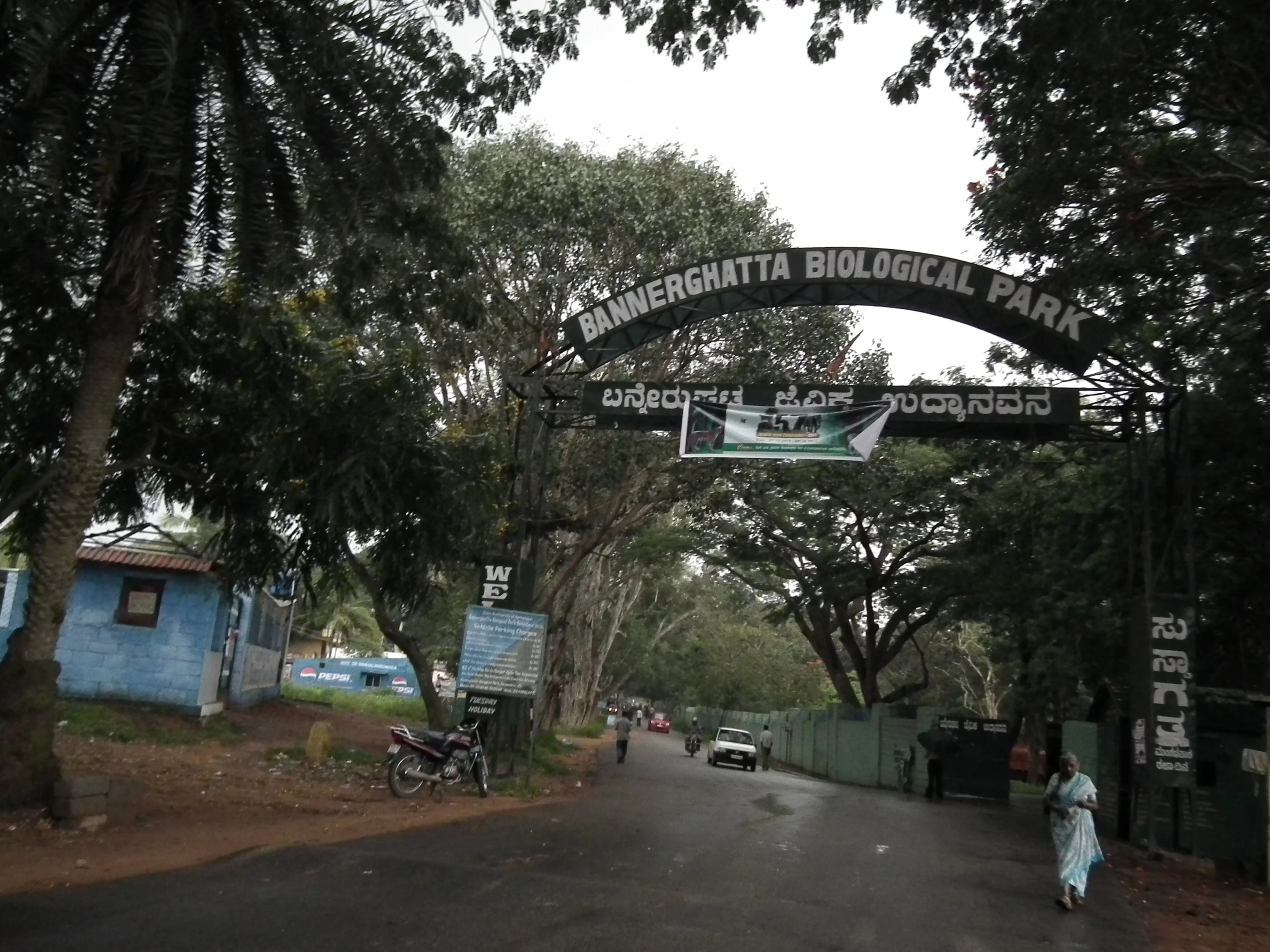 Entrance sign for Bannerghatta Biological Park with text in English and Kannada, road leading into park, trees and buildings visible