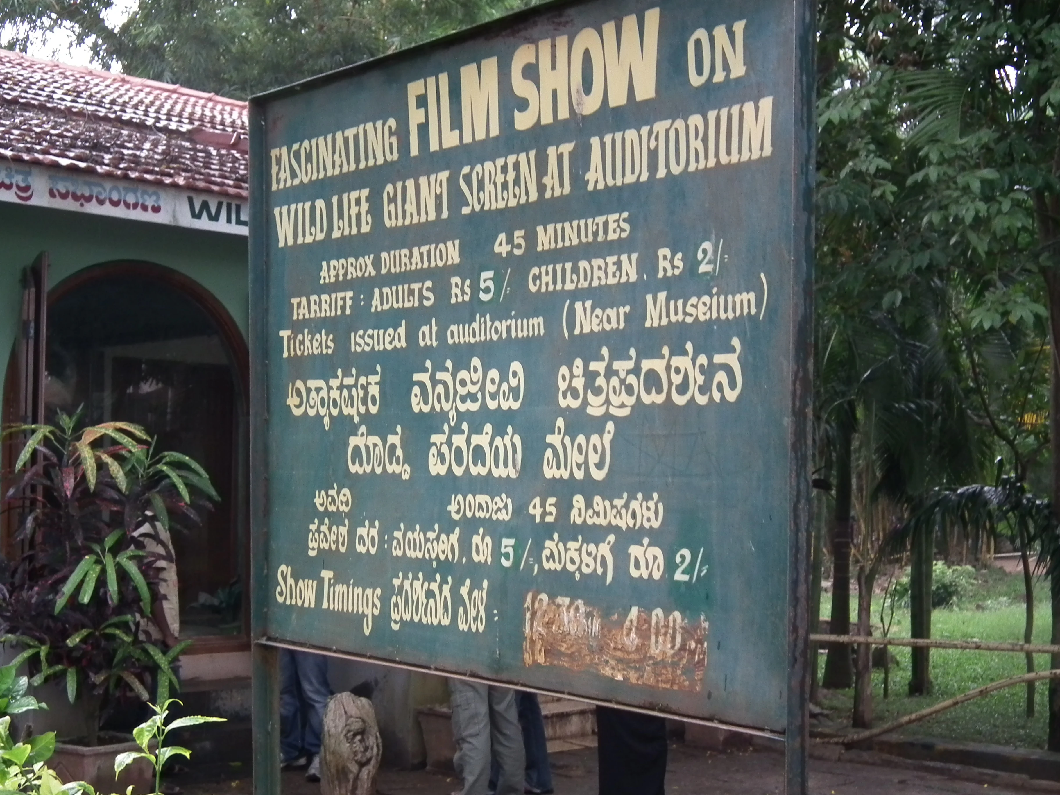 Green signboard with text about wildlife film show at auditorium, showing ticket prices and show details in English and Kannada