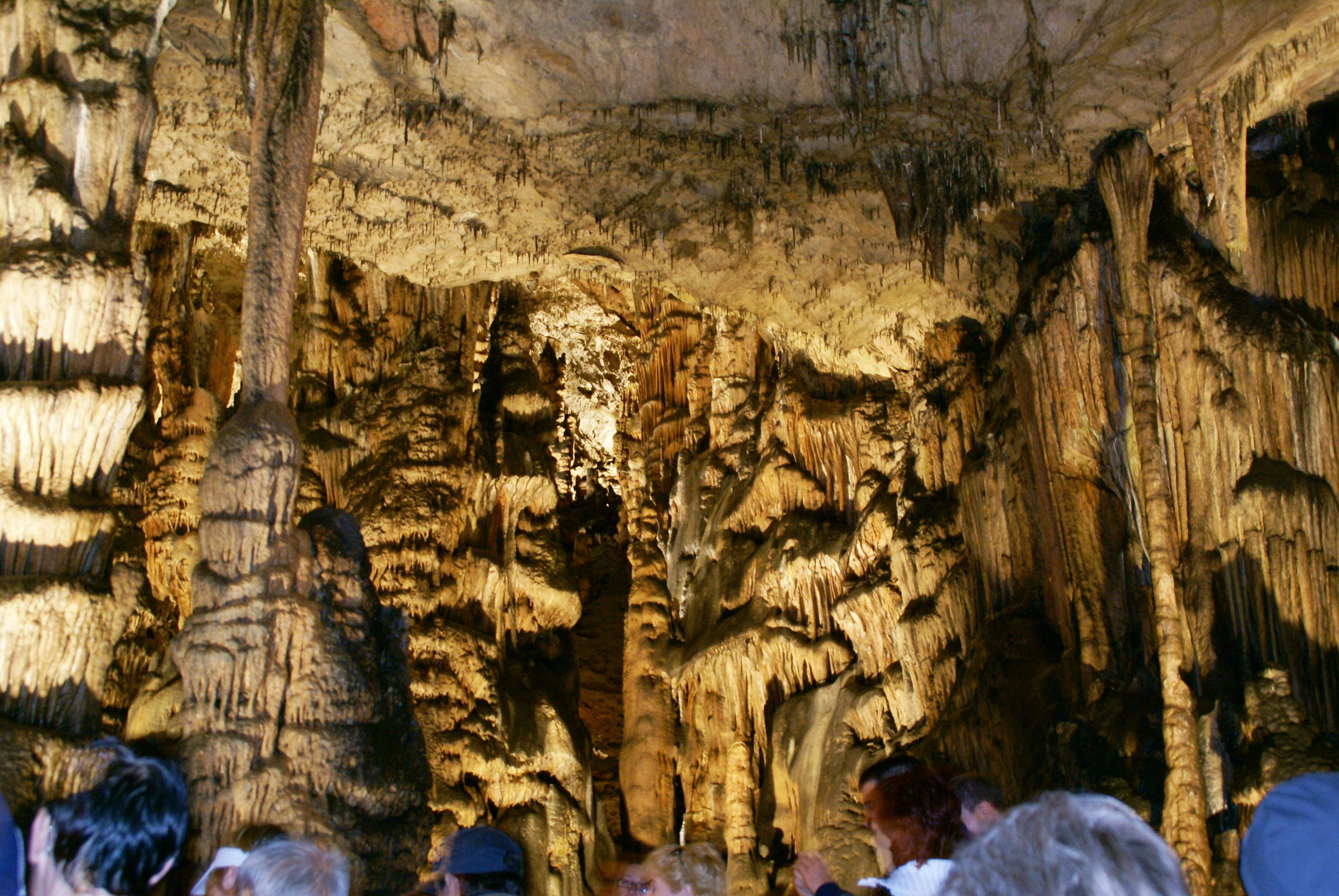 A view inside a cavern showing large dripstone formations, stalactites, and stalagmites under warm artificial lighting, with the tops of people's heads visible at the bottom of the frame.