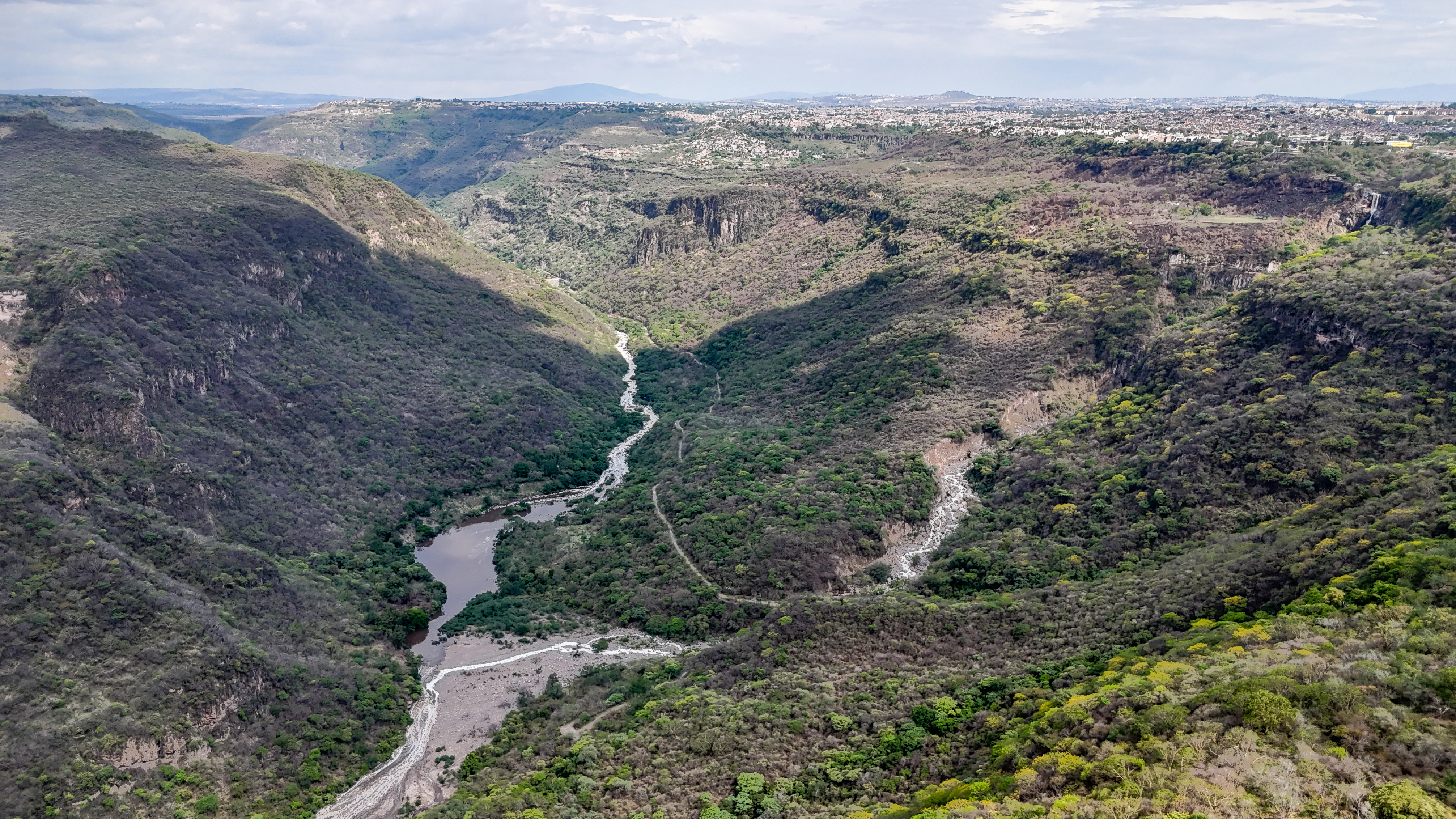 An aerial drone shot looking down into the deep, steep-walled canyon of Barranca de Oblatos, with the winding Santiago River flowing through the bottom and the surrounding landscape covered in scrubby forest and canyon vegetation.